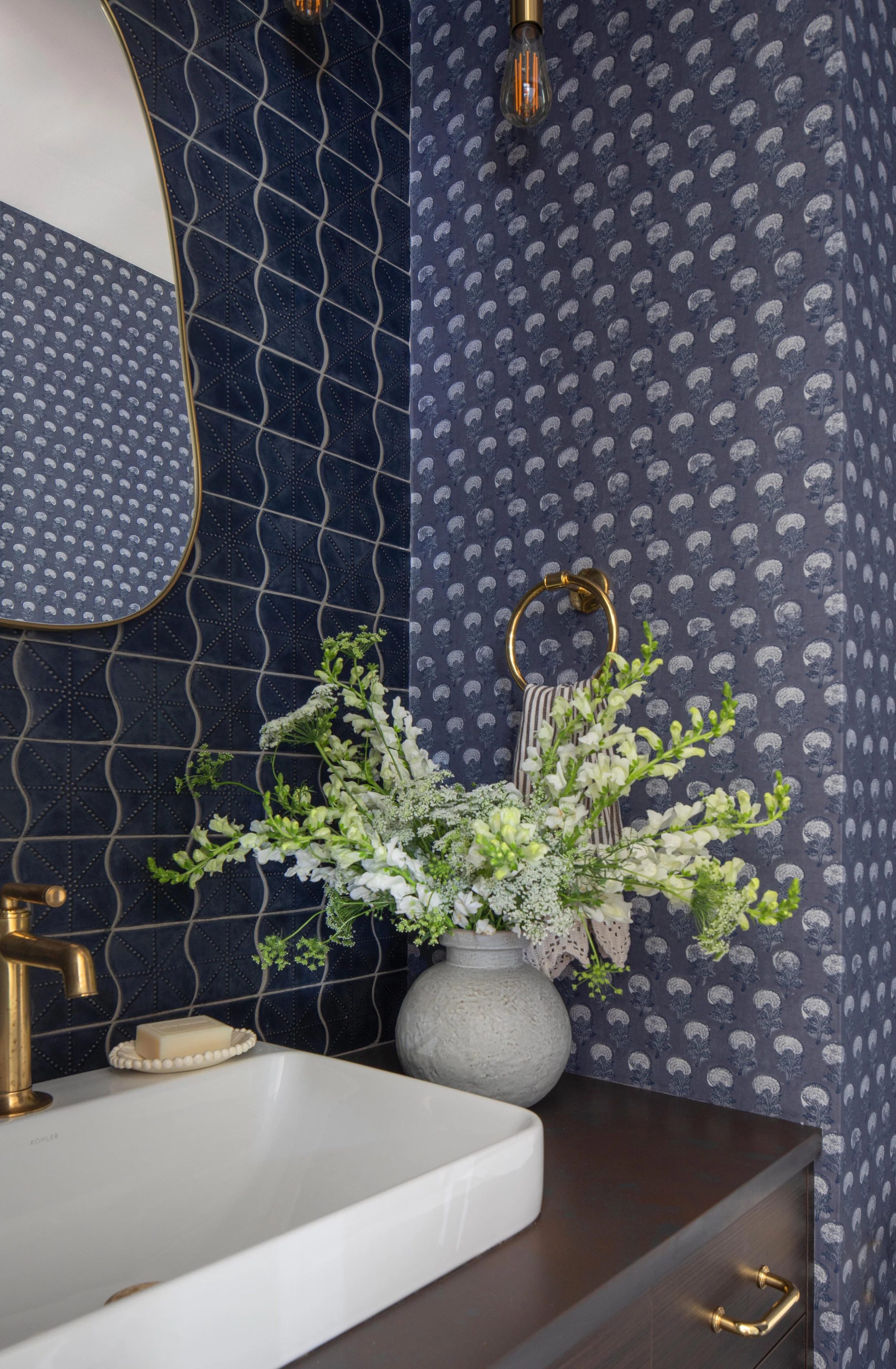 Bathroom sink with a soap dish and bar of soap, a large white ceramic basin, a gold faucet, a dark wooden vanity, a large vase with white and green flowers, a navy blue patterned wall with two different wallpaper designs, a mirror with a gold frame, 