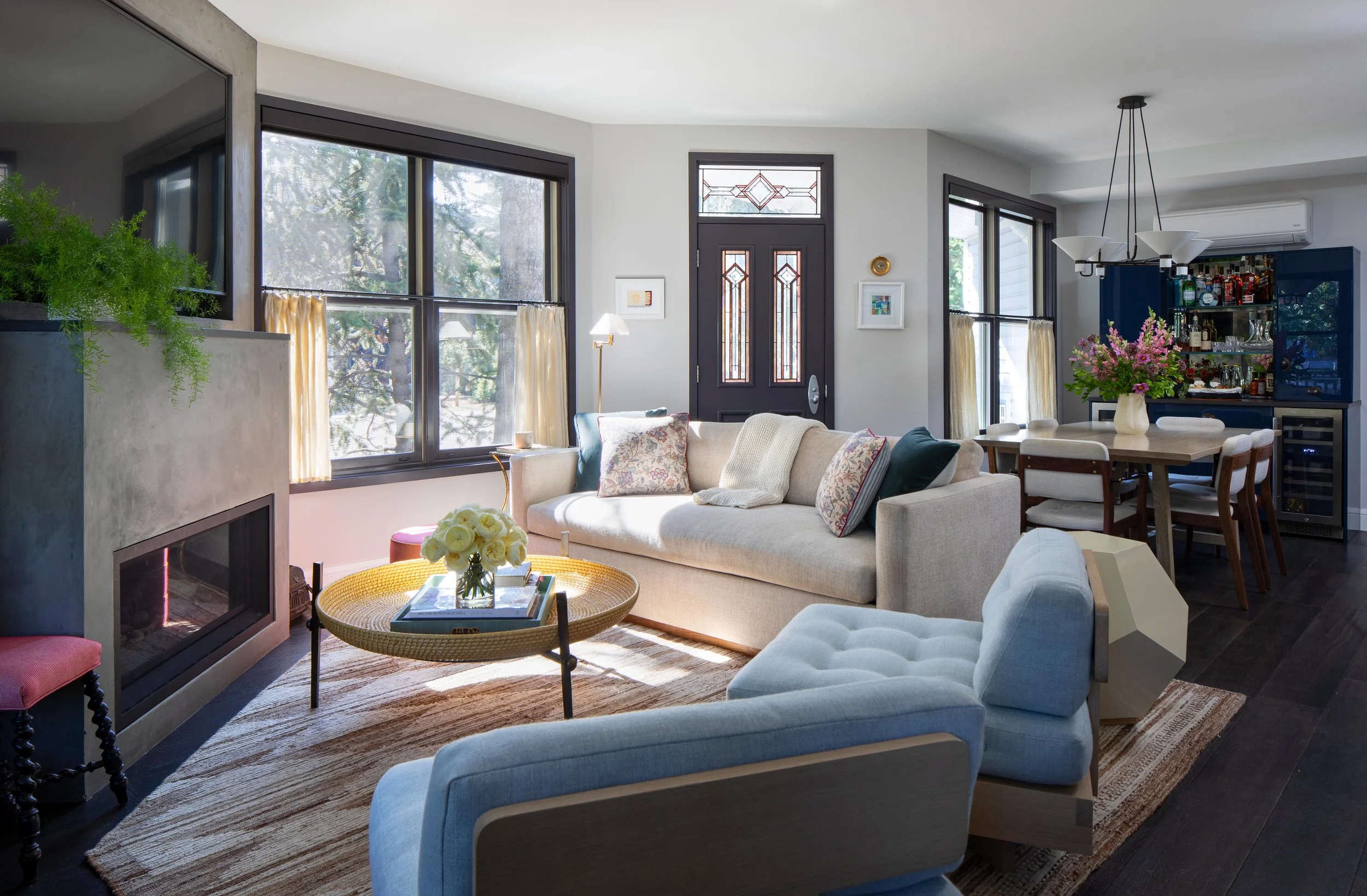 Bright living room with large windows, beige sofa, blue armchair, fireplace, round coffee table with flowers, wooden dining table with chairs, bar area with bottles, and dark wood flooring.