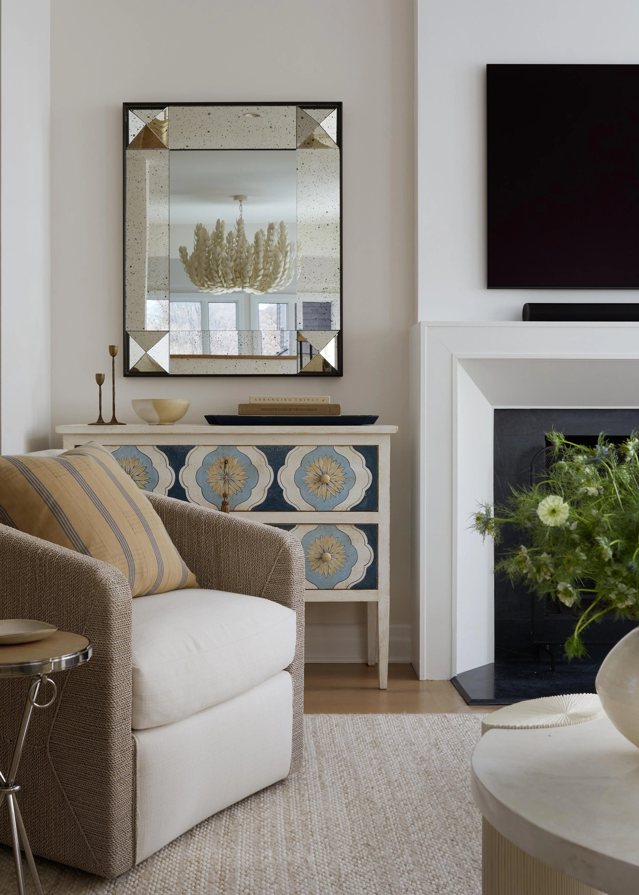 Living room corner with a beige sofa, striped pillow, ornate blue and white patterned cabinet, large mirror reflecting a chandelier, and a white fireplace with a black mount, decorated with books and candlesticks, and a bouquet of white flowers.