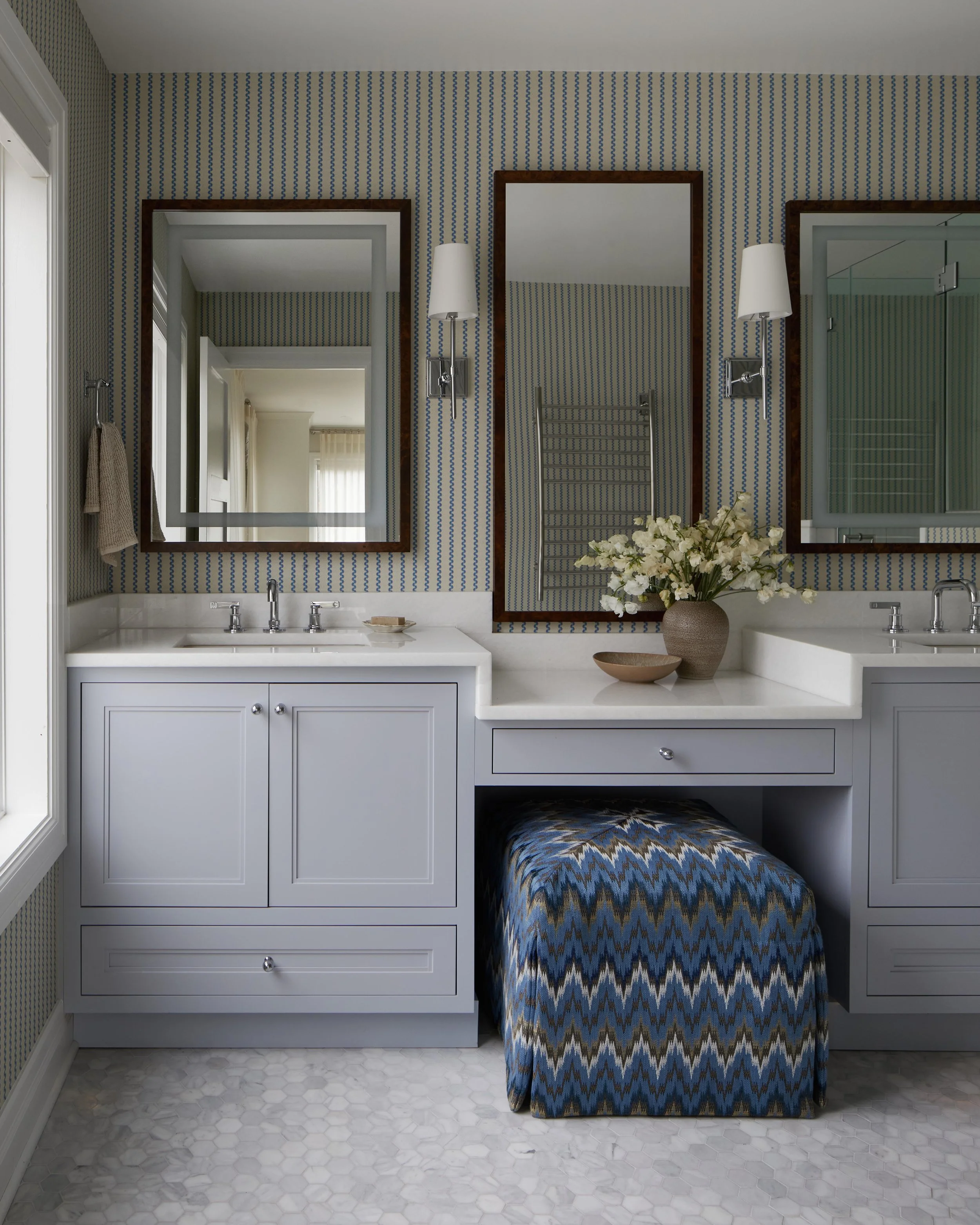 Bathroom with double vanity, mirrors, wall-mounted lamps, floral arrangement, and patterned ottoman.
