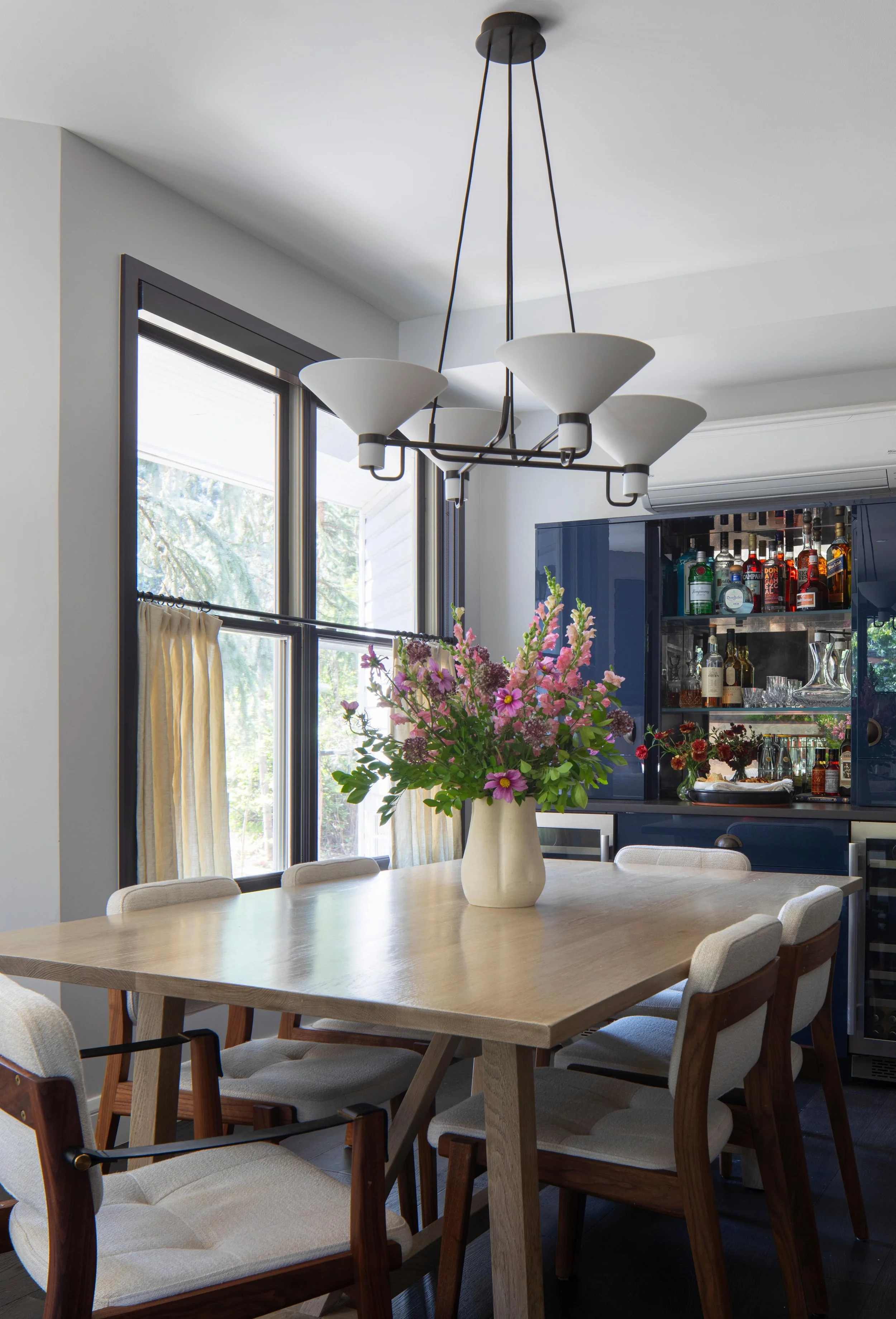 A dining room with a wooden table, eight upholstered chairs, a large ceramic vase filled with pink and purple flowers, a navy blue china cabinet, and a window with light yellow curtains. There is a modern chandelier hanging above the table.