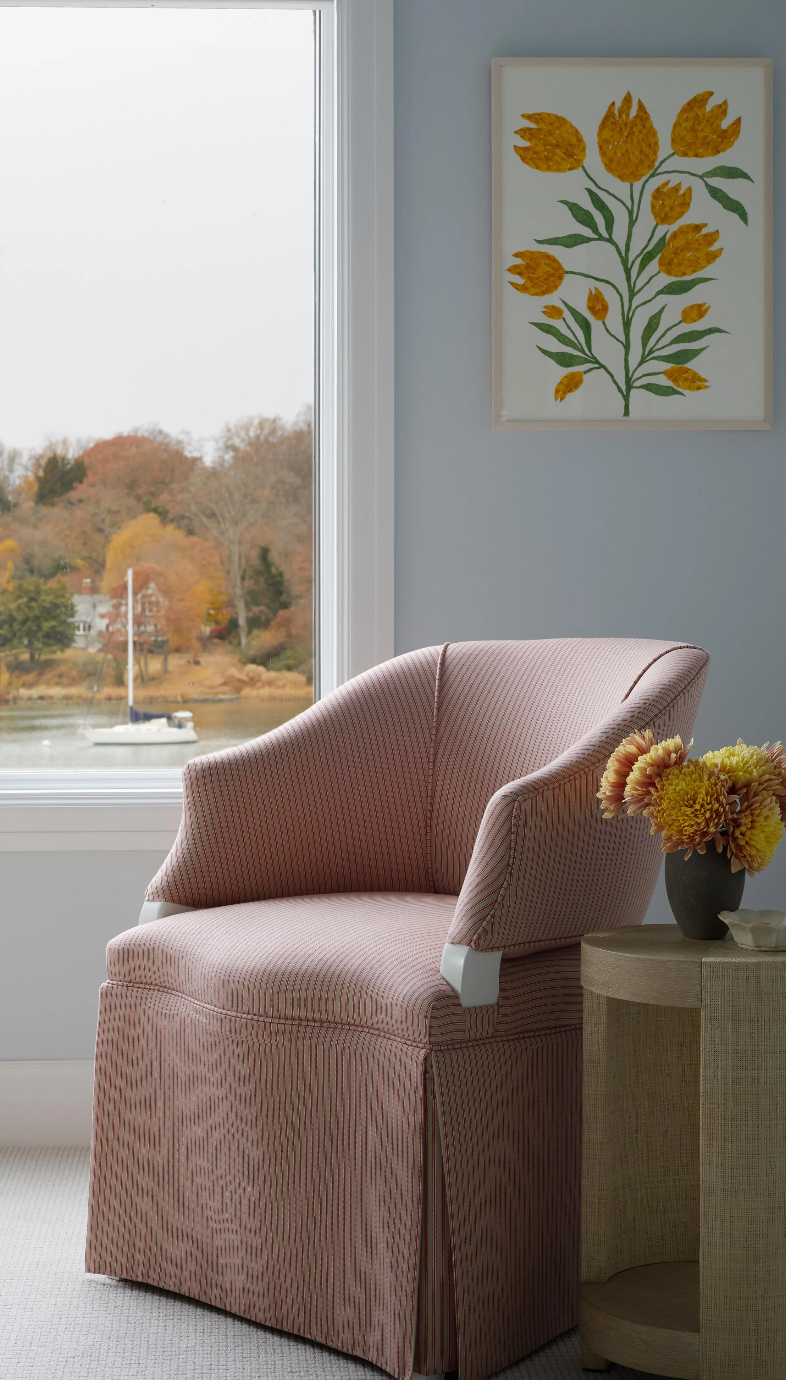 A cozy interior corner with a pink striped armchair, a wooden side table with a dark vase filled with yellow and orange flowers, and a framed botanical art of yellow flowers on the wall. Outside the window, fall foliage and a sailboat on a lake are v