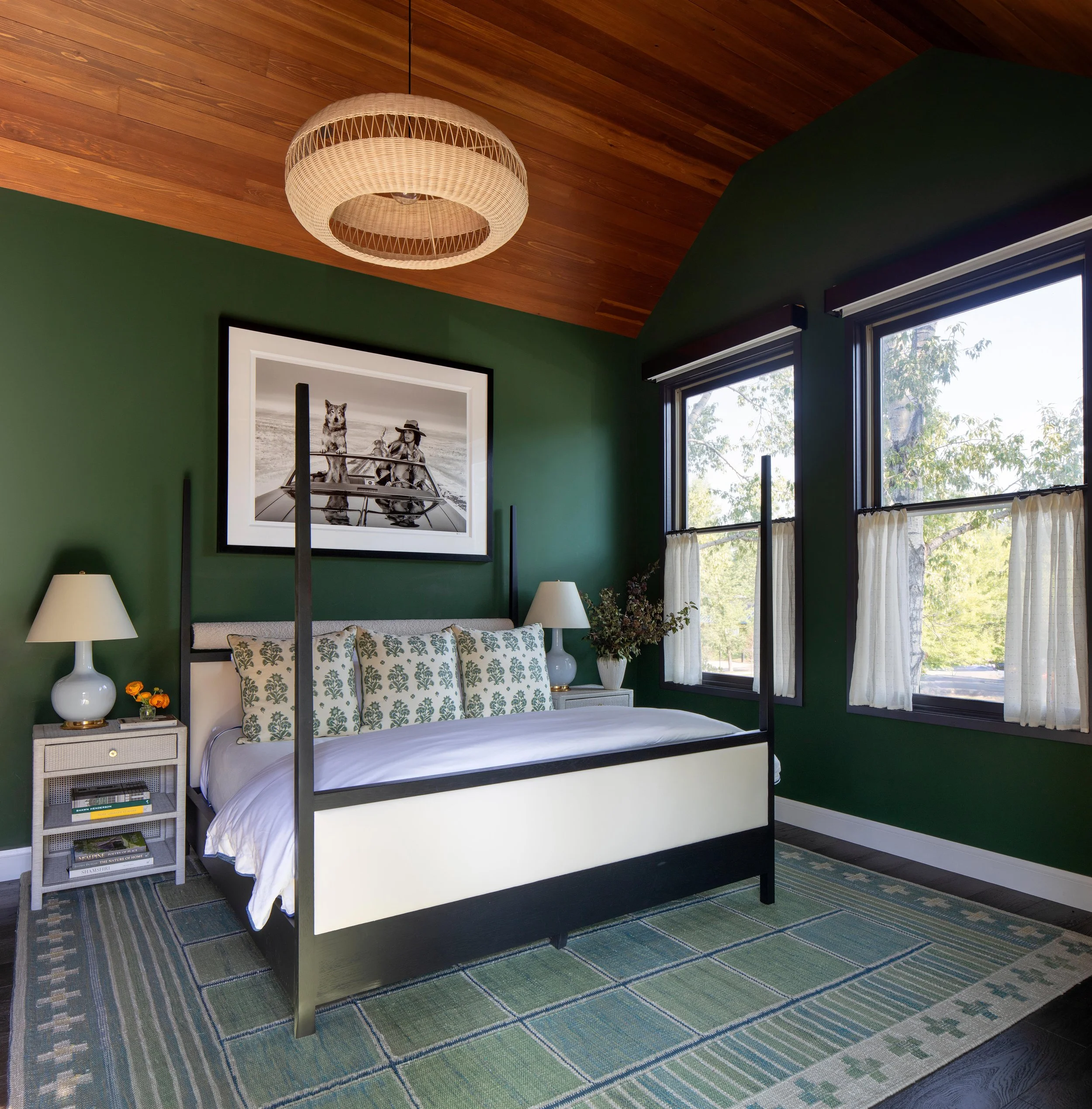 A bedroom with dark green walls, a white bed with black frame, green patterned pillows, two white nightstands with lamps, a framed black-and-white photograph of a dog and person on a boat, and two windows with white curtains showing trees outside.