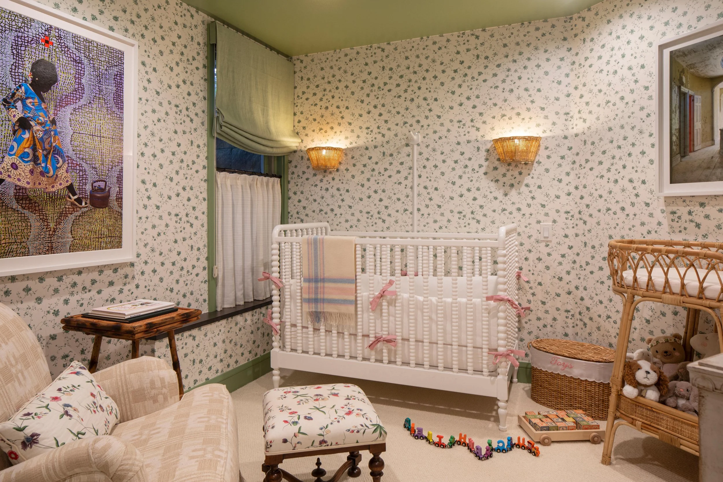 A cozy nursery with floral wallpaper, a white crib decorated with pink bows, a rattan changing table with stuffed animals, a floral upholstered ottoman, a floral sofa with a pillow, a small wooden side table, colorful toy trains on the floor, a frame