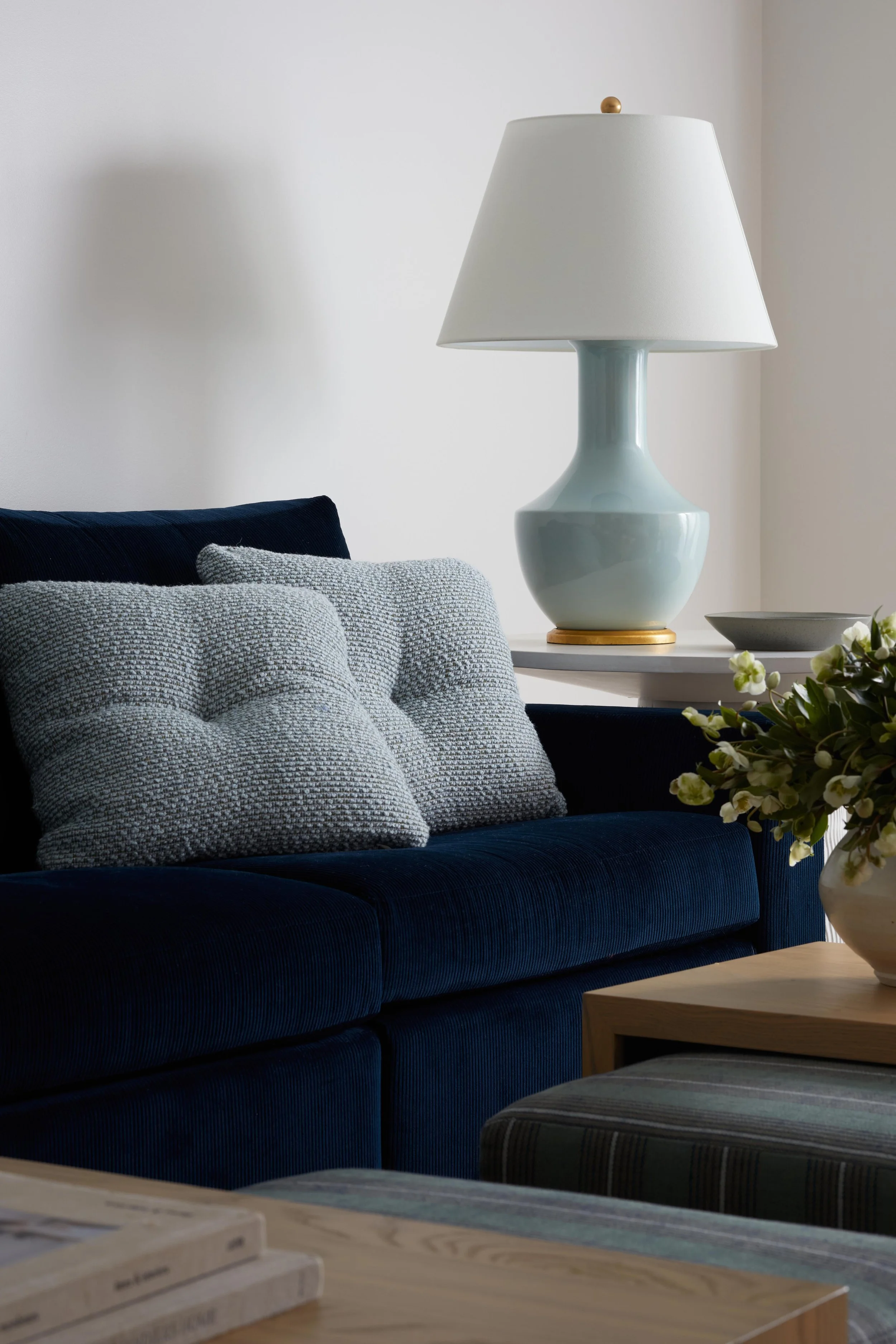 Interior of a living room with a navy blue sofa, a light gray textured pillow, a side table with a white lamp, a gray bowl, and a vase with white flowers.