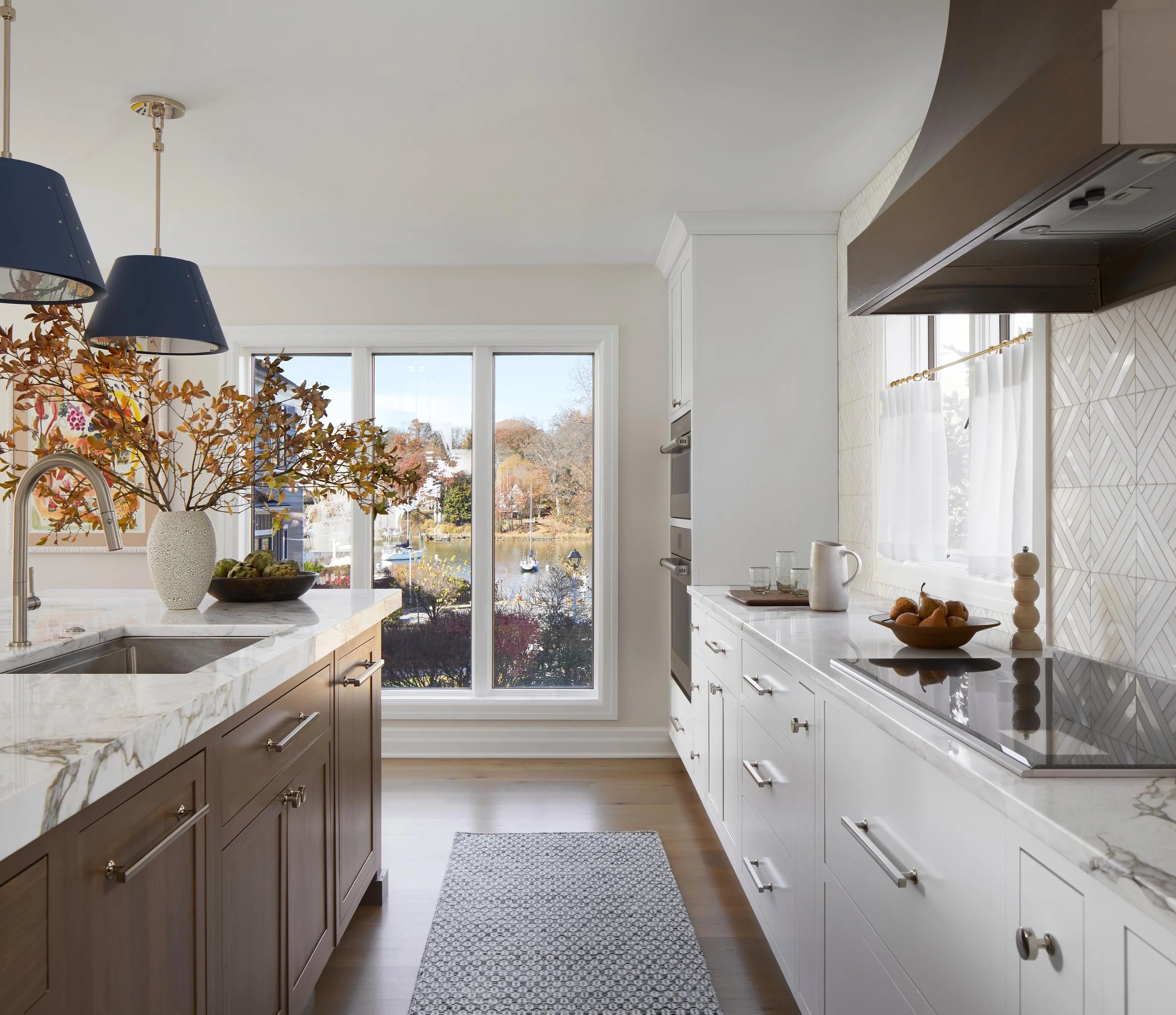 Bright kitchen with white cabinets, marble countertops, large window overlooking a lake, decorative vase with flowers, and a bowl of fruit.