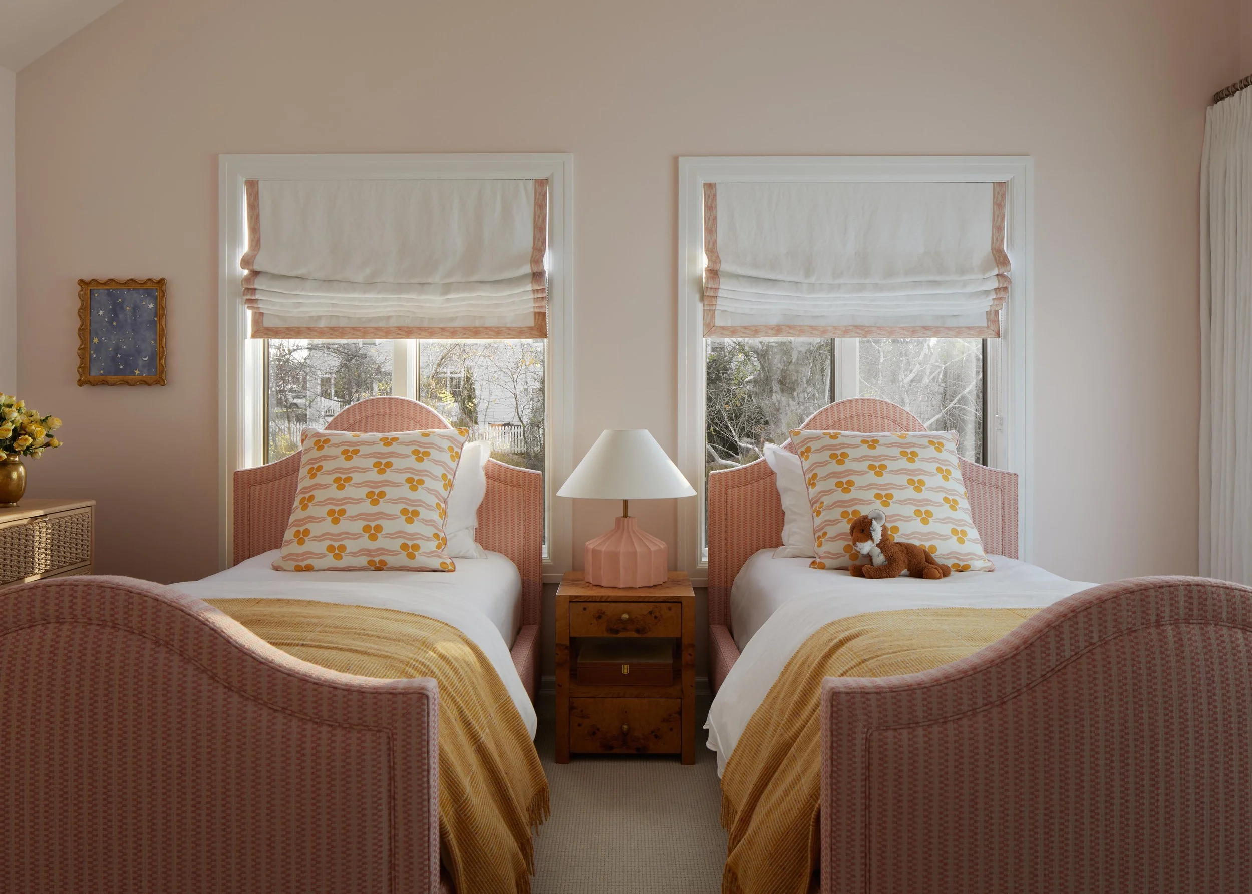 A bedroom with two twin beds, each with pink headboards, white bedding, yellow blankets, and pillows with yellow flower patterns. A plush teddy bear sits on the right bed. Between the beds, there is a wooden nightstand with a pink lamp. Behind the be