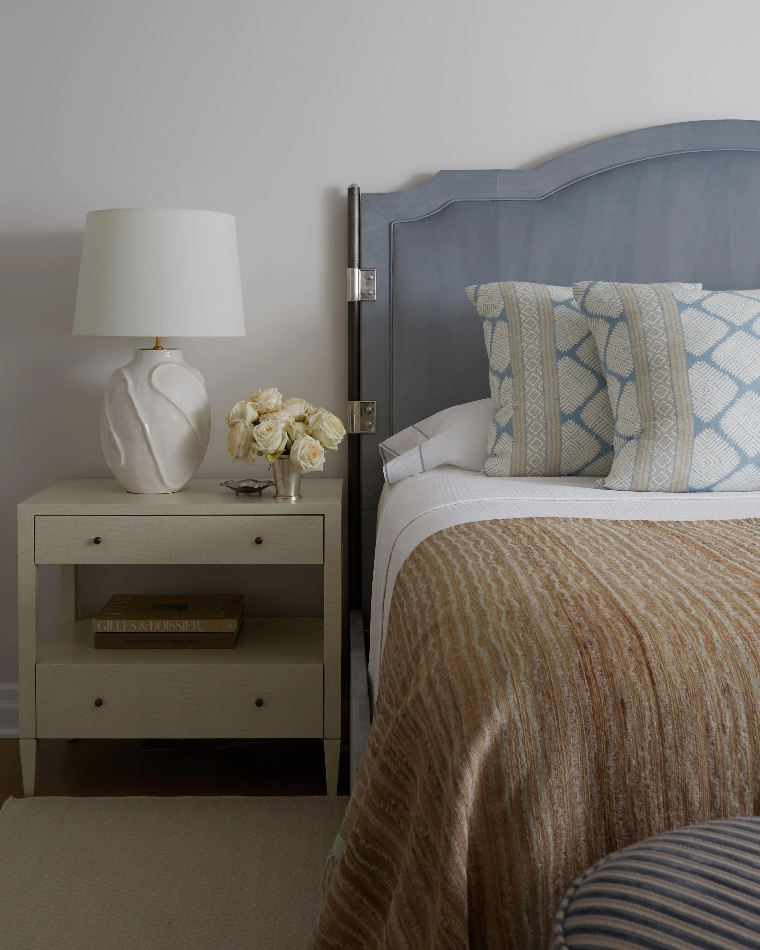 Close-up of a bedroom corner with a white nightstand, stacked books, a table lamp, a flower vase, a bed with patterned pillows, a headboard, and a beige bedspread.
