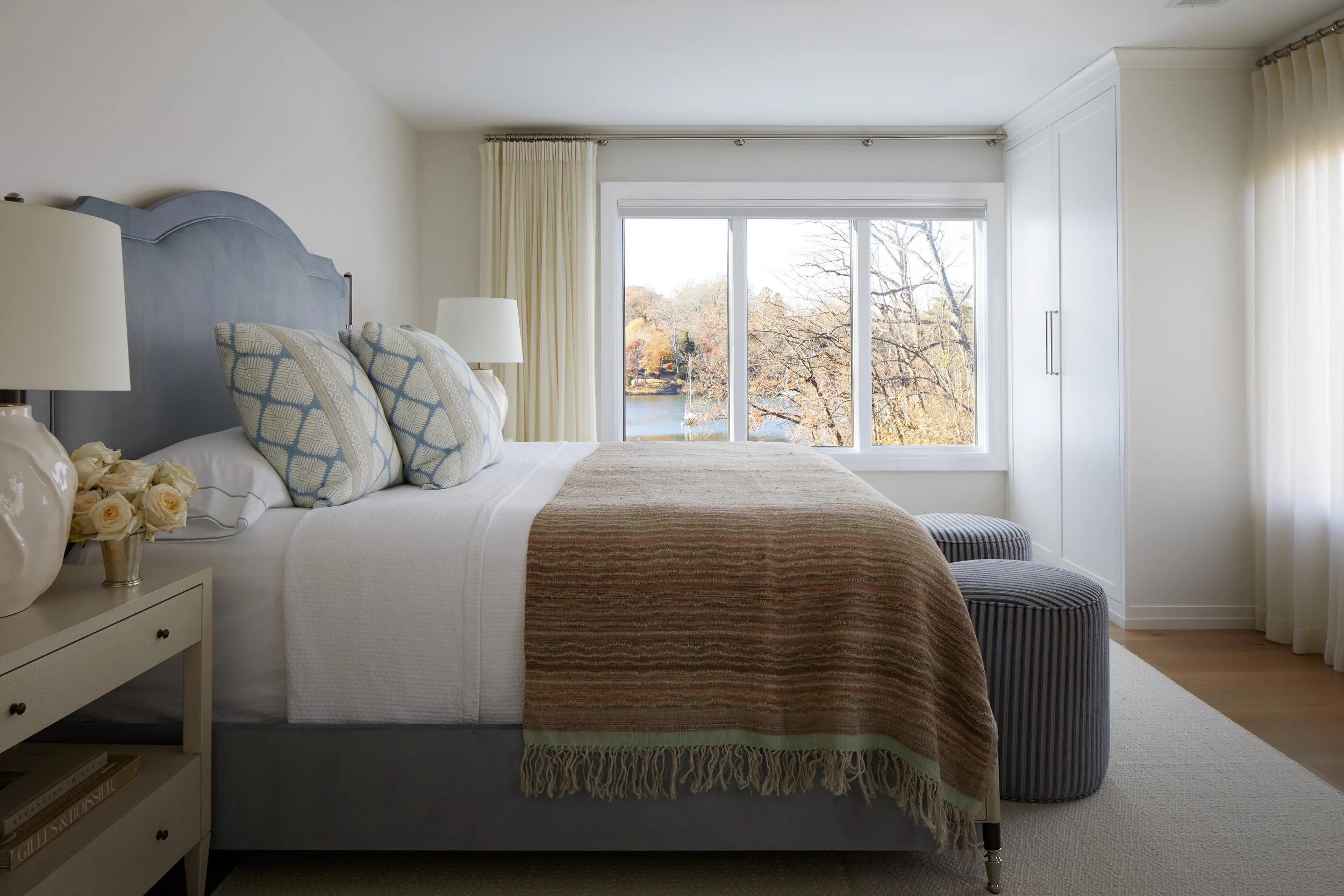Modern bedroom with large window overlooking trees and water, featuring a bed with white linens, patterned pillows, bedside lamps, a stripe ottoman, and a white wardrobe.
