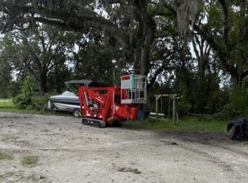 A red cherry picker lift with a basket paused outdoors near trees and a white boat on a trailer.