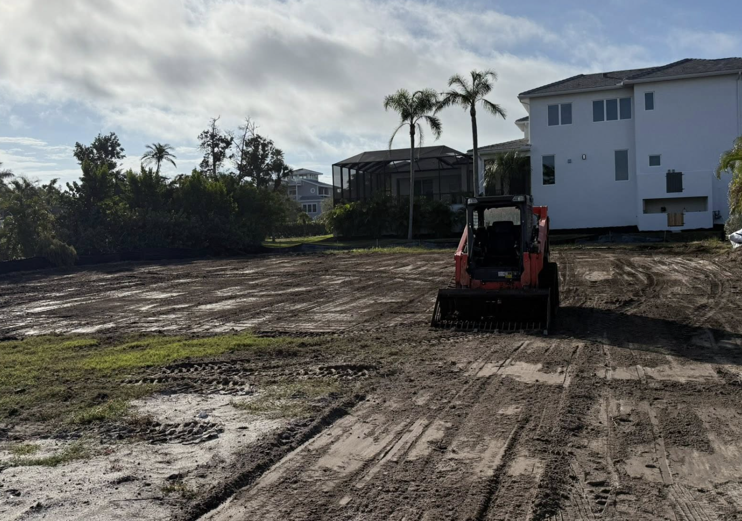 A construction site with a small red skid-steer loader on bare dirt, near a new white house with a screened porch and palm trees in the background.