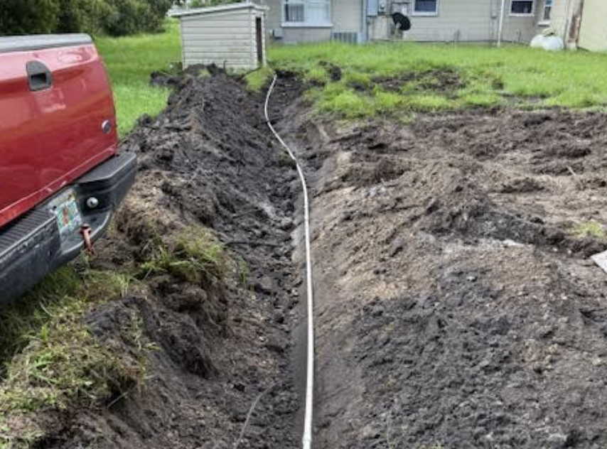 A trench dug in the ground with a white pipe running through it, next to a red truck and a grassy area, with a house and shed in the background.