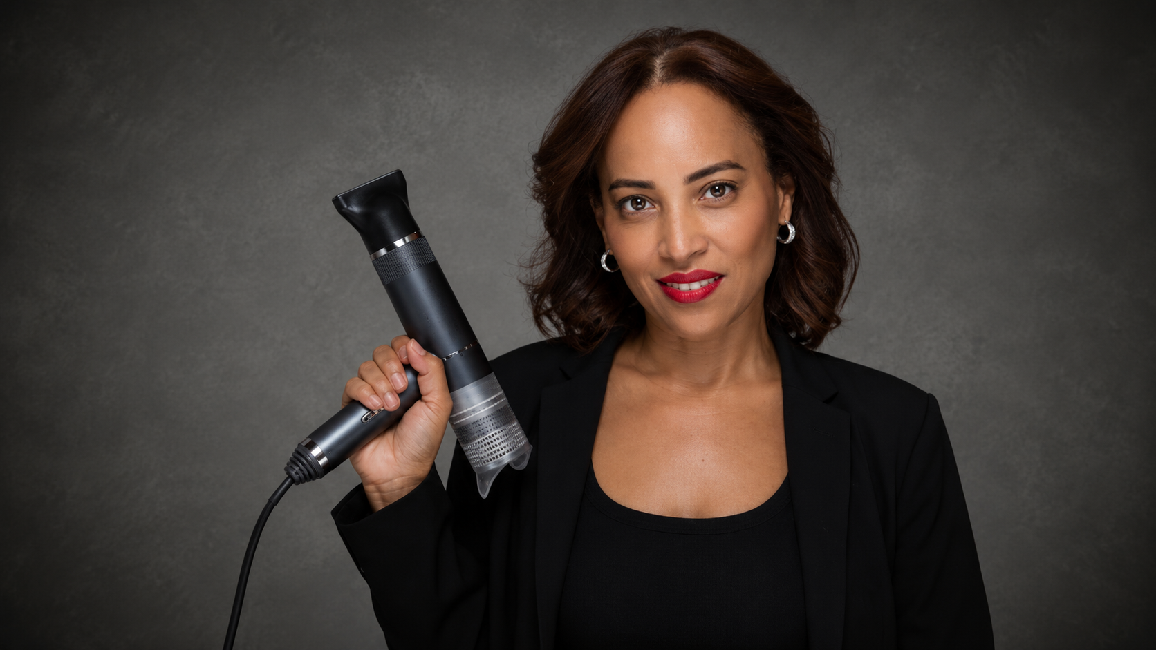 A woman with shoulder-length brown hair, wearing a black blazer and top, holding a round hairbrush and a blow dryer against a textured gray background.