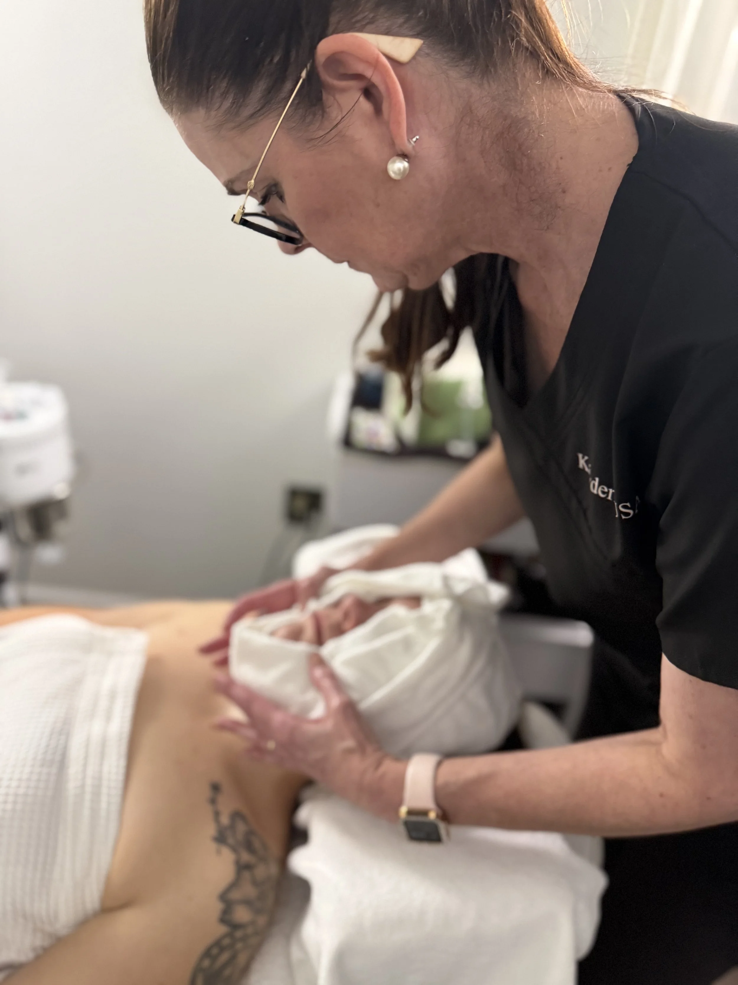 Close-up of a woman with glasses and pearl earrings, performing a medical procedure on a patient's hand, in a clinical setting.