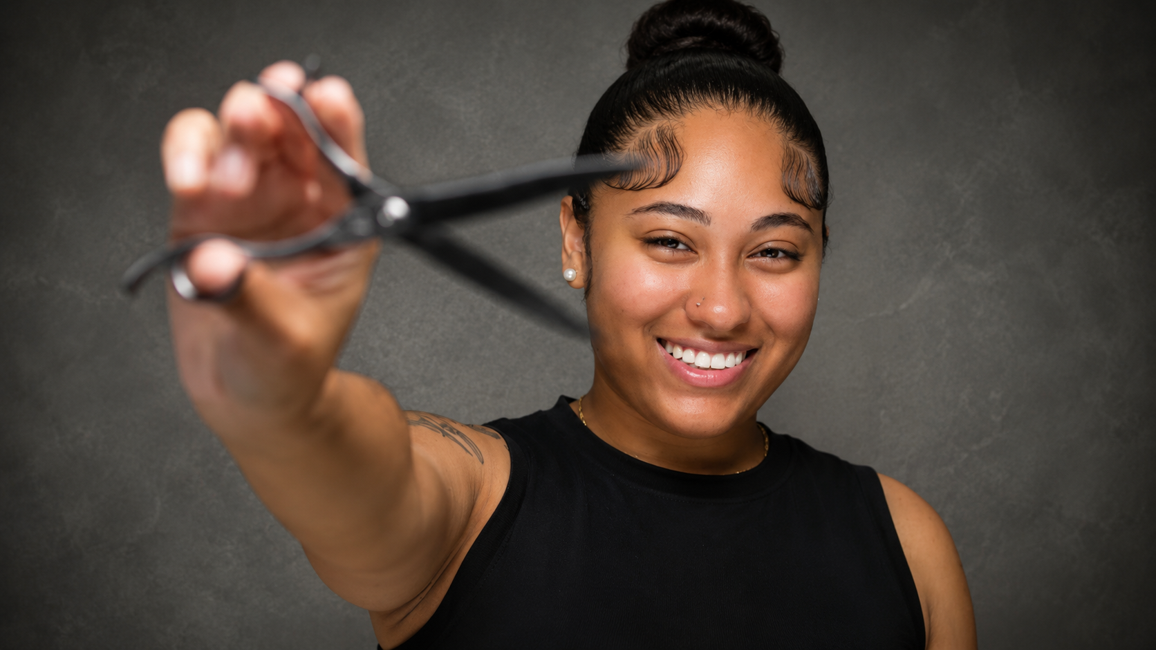 A woman with a bun hairstyle, smiling and holding a pair of hair scissors towards the camera, wearing a black sleeveless top, standing against a gray background.