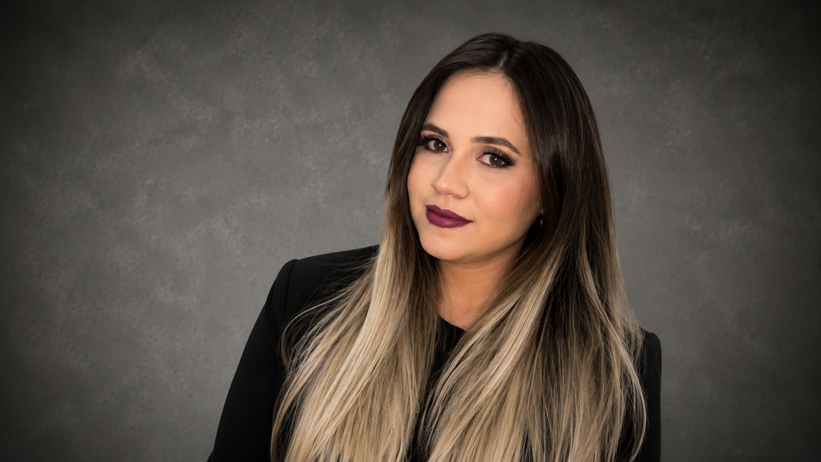Portrait of a woman with long blonde hair, wearing a black blazer, and makeup with dark lipstick, posed against a gray background.