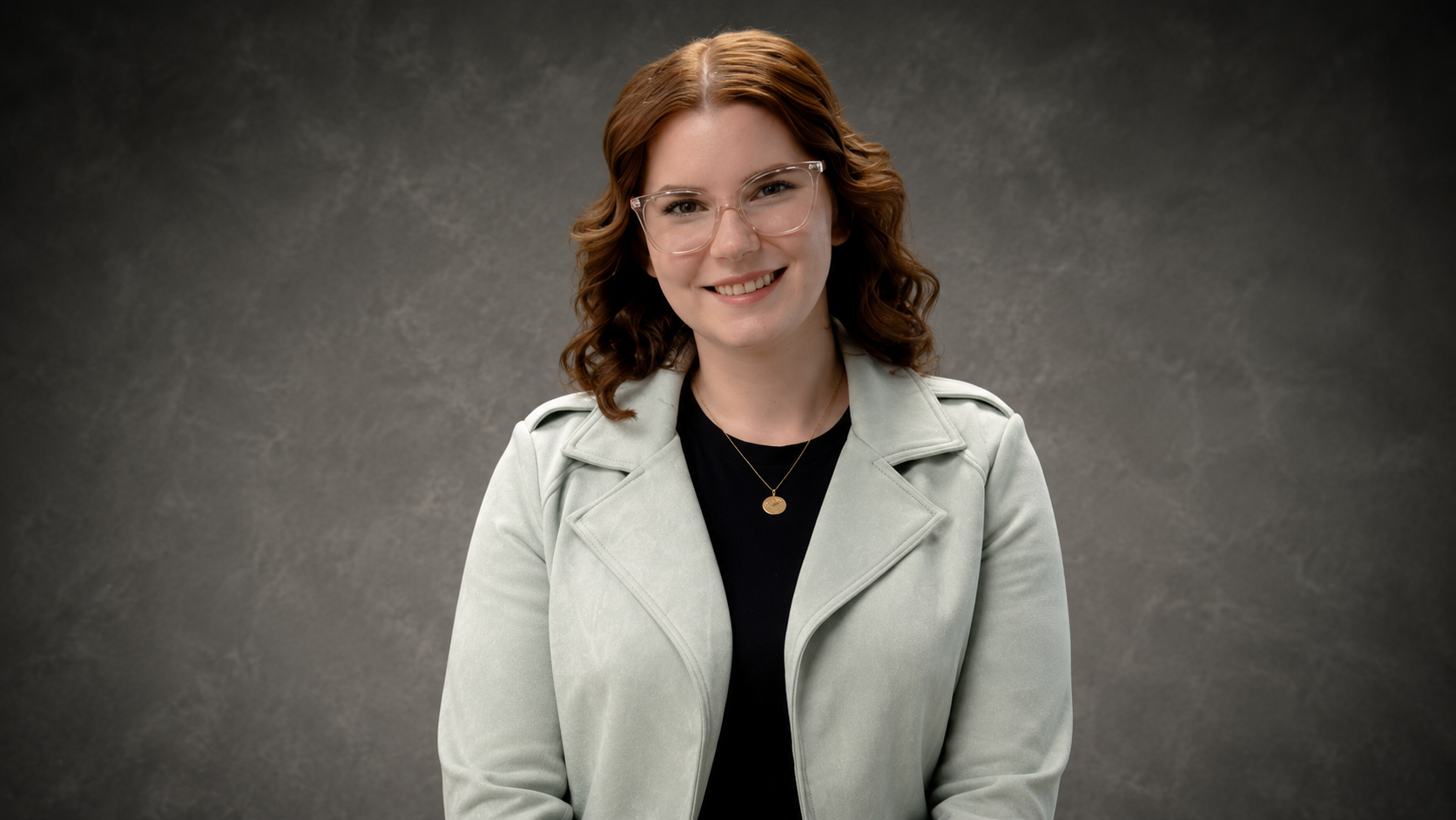 A smiling woman with shoulder-length wavy red hair, wearing clear glasses, a black top, a light gray jacket, and a gold necklace, posing against a dark gray textured background.