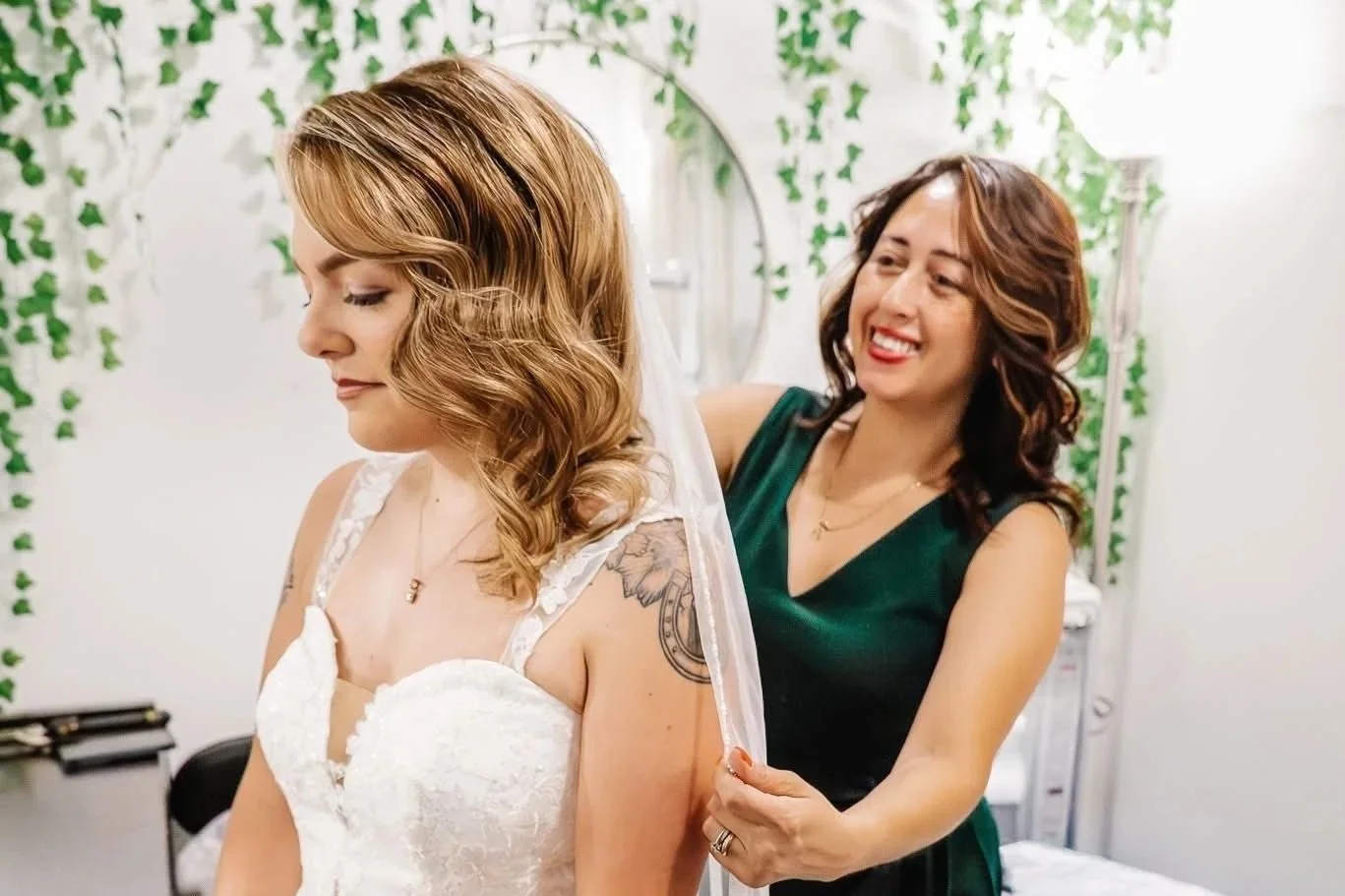 A woman with wavy brown hair is helping a bride with her veil in a decorated room. The bride has light brown hair and is wearing a white lace wedding dress. The woman assisting her is smiling and wearing a dark green dress.