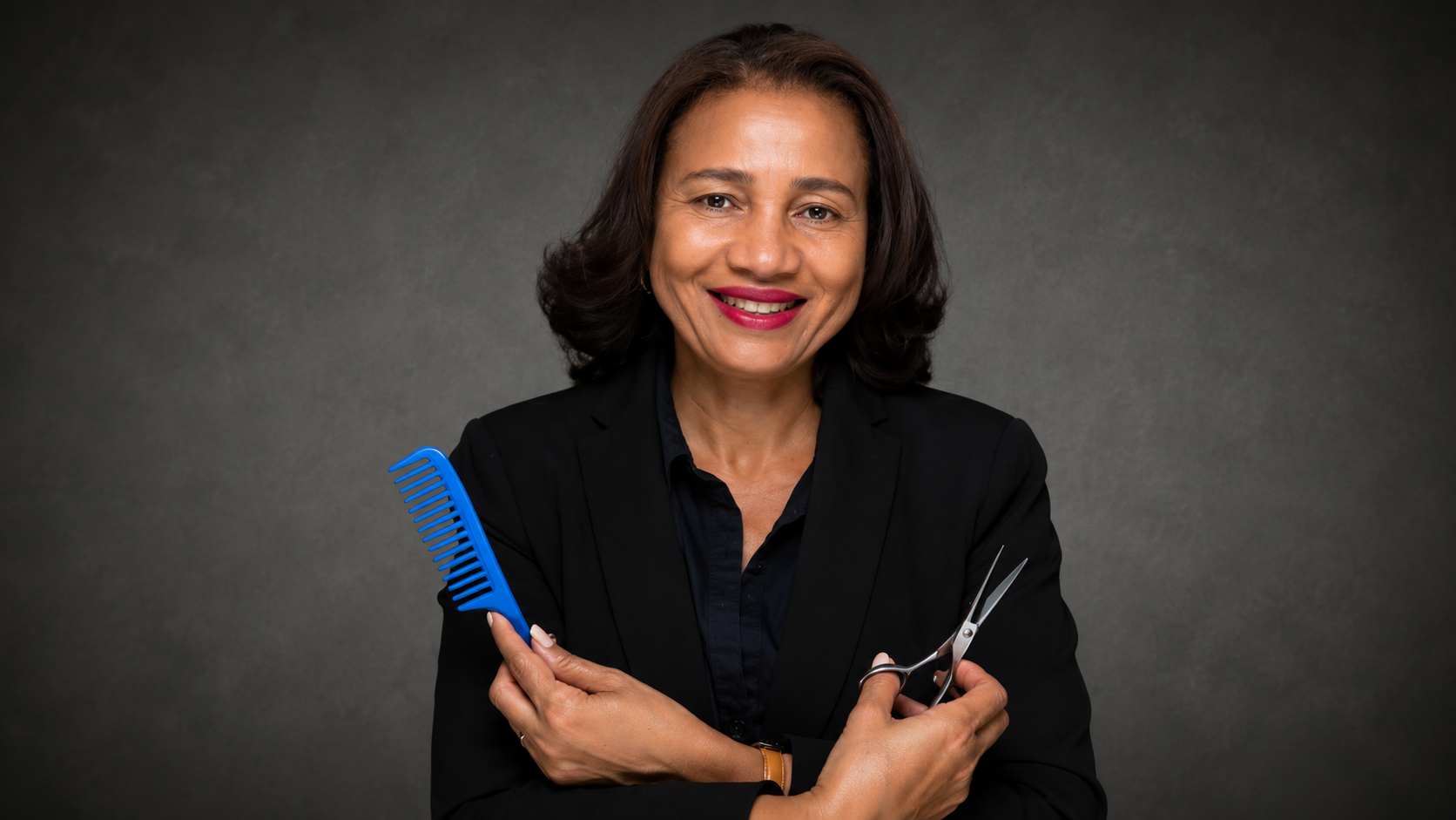 A woman with shoulder-length dark hair, wearing a black blazer and shirt, holding a blue comb in one hand and a pair of scissors in the other, smiling against a gray background.