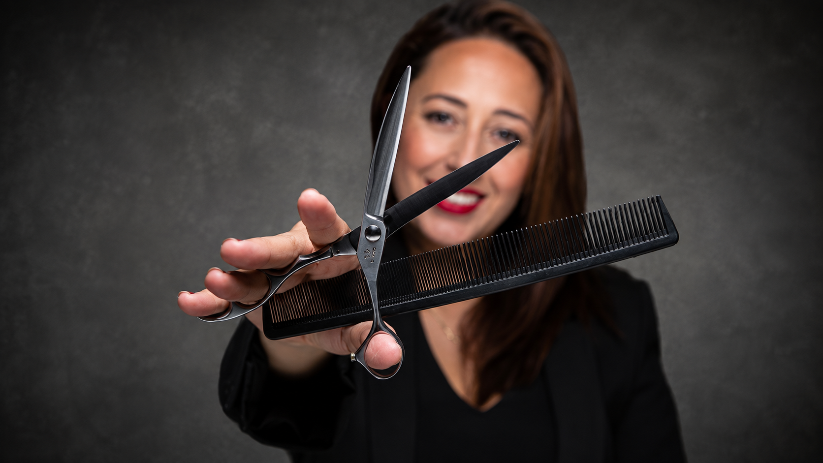 A woman smiling, holding a pair of scissors and a large black comb, with a dark gray background.