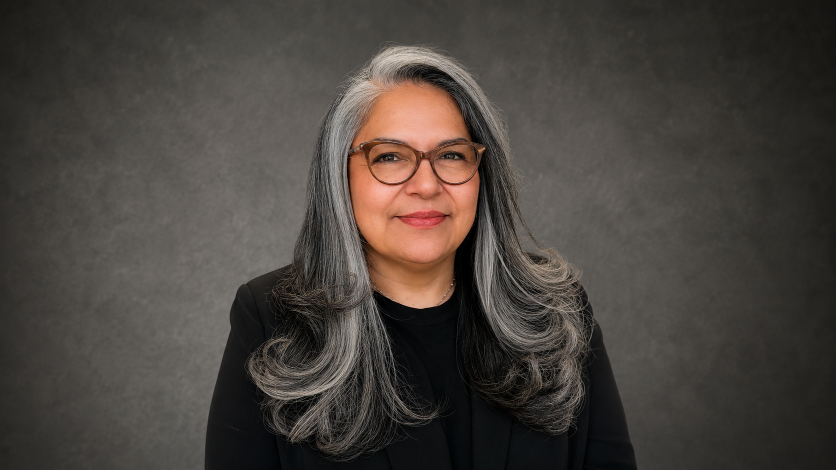 A woman with long gray hair, glasses, and a black blazer smiling in front of a gray background.