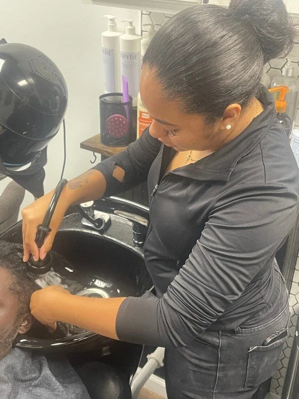 A hairstylist rinsing a client's hair in a black salon sink, with hair care products on a shelf behind her.