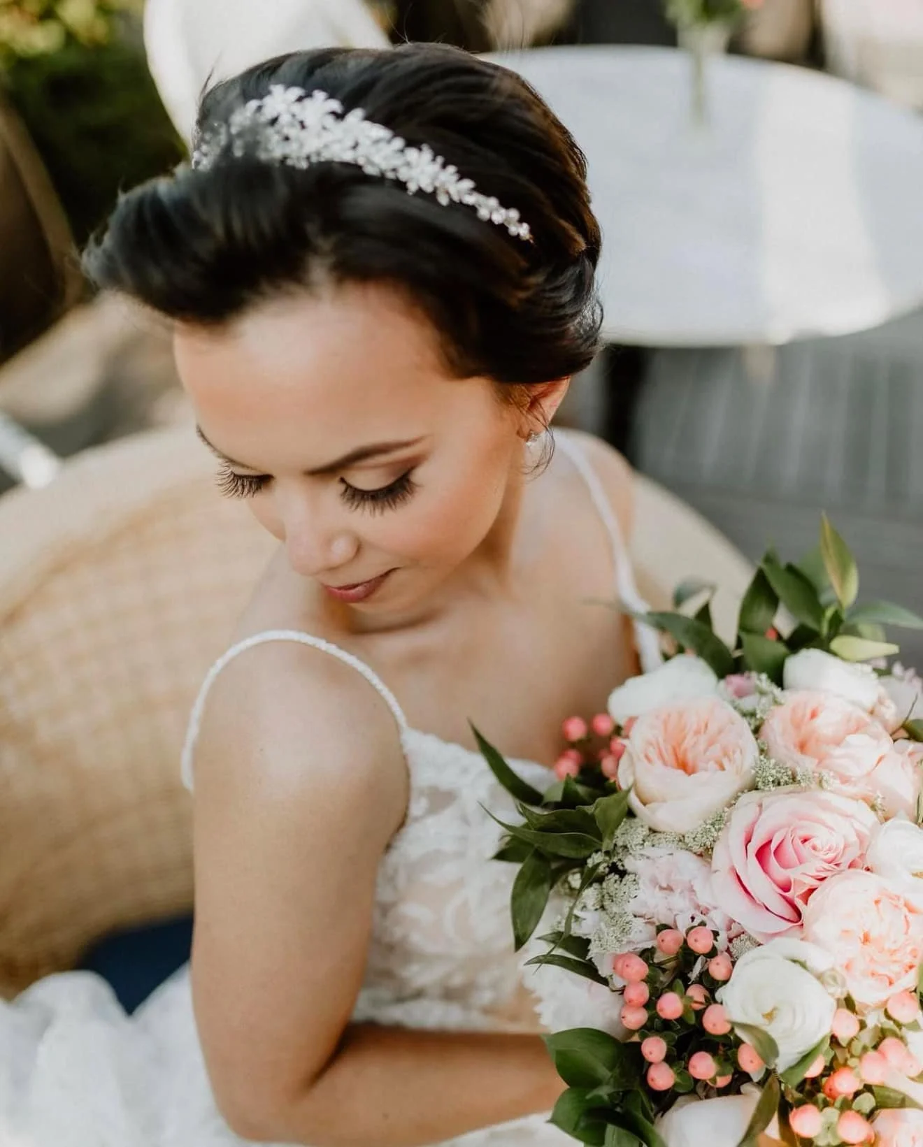 A bride with dark hair and a floral headband, wearing a white wedding dress, holding a bouquet of pink and white roses, sitting on a beige armchair.