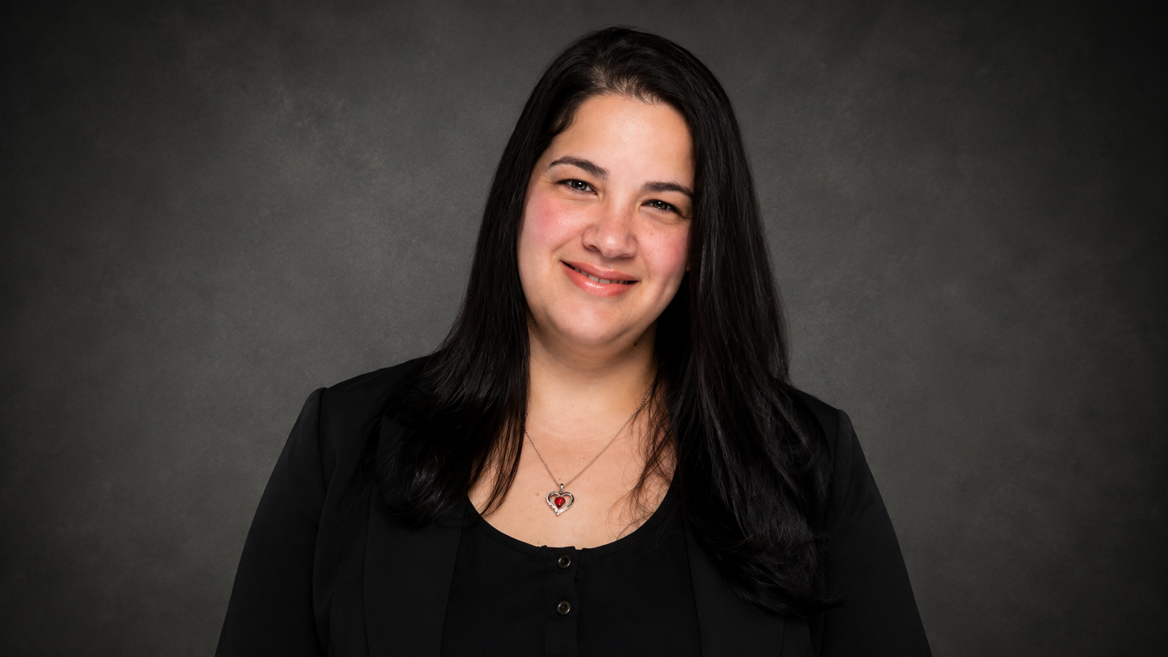 A woman with long dark hair, wearing a black top and blazer, smiling, with a gray background.