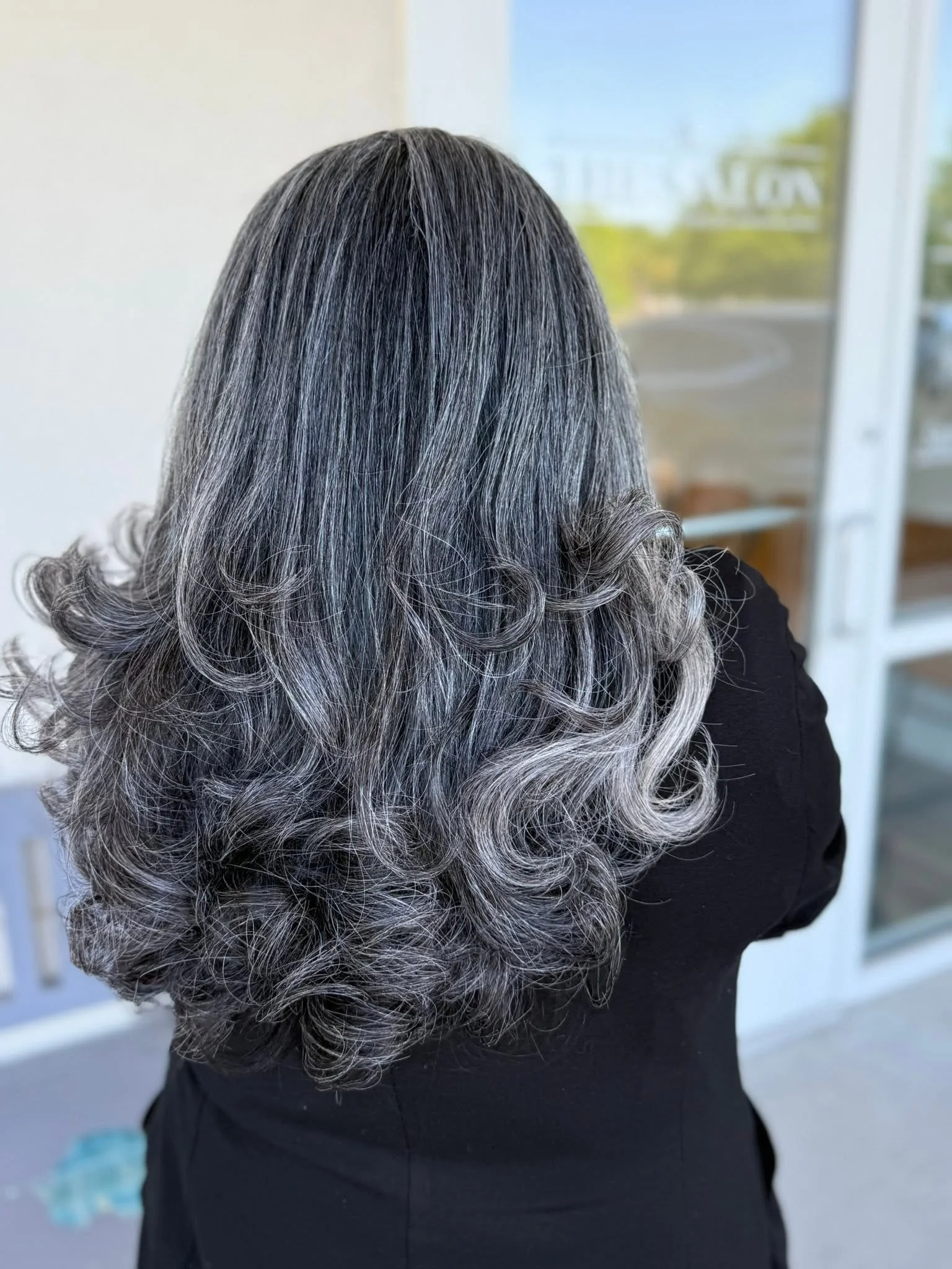 A woman with long, curly gray hair standing outdoors near a window or glass door.