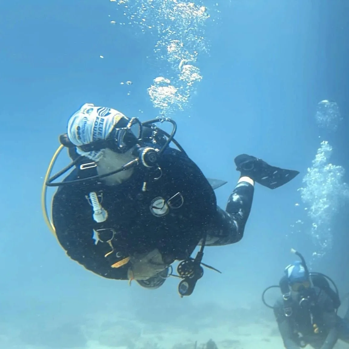 Two scuba divers underwater with one closer to the camera and bubbles rising above. The closer diver is  wearing full scuba gear, including mask, regulator, and fins.