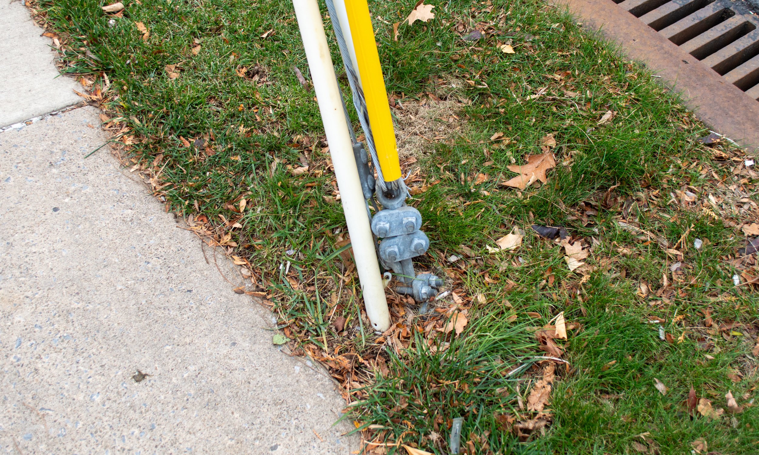 Utility pole guy wire anchor grounding point with yellow safety casing and metal tension hardware next to sidewalk.