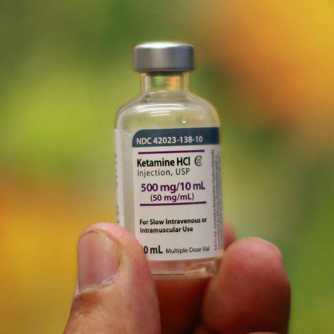 Close-up of a small medical vial labeled 'Ketamine HCl' being held between a person's thumb and index finger. The background is blurred with green and yellow tones.