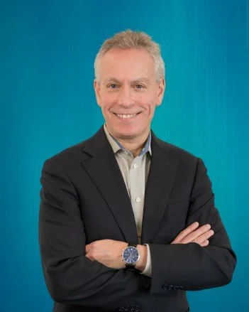 A smiling man with blonde hair, wearing a black blazer, light-colored shirt, and a watch, standing against a blue background with his arms crossed.
