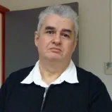 Un homme âgé avec des cheveux gris, portant une chemise blanche et un pull noir, se trouve devant un mur beige dans une pièce intérieure.