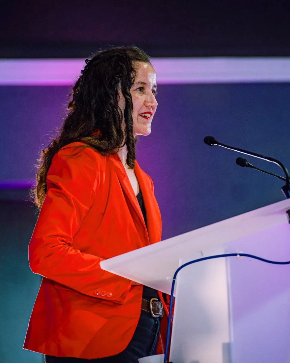 Photo de la candidate LFI Valentine Mercier en meeting portant une veste rouge. Elle est de 3/4 et regarde à droite de la photo. Devant elle un pupitre blanc avec deux microphones, dans une salle bien éclairée.
