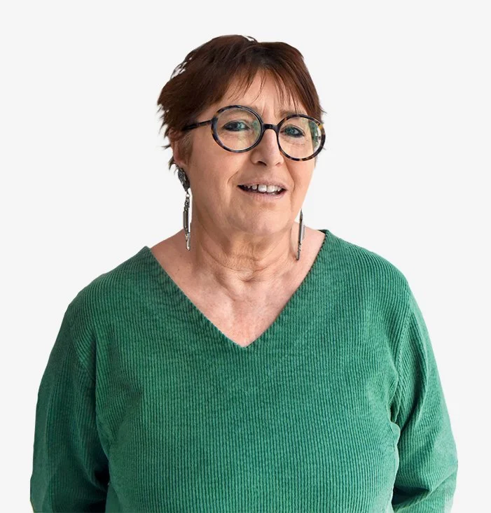 Photographie de face d'une femme qui souri la bouche ouverte. Elle a les cheveux courts châtains dans les tons rouge-orangés. Elle porte des lunettes rondes, des boucles d'oreille longues et un pull vert à coll V.