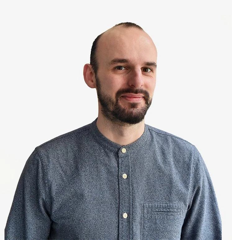 Homme de face, souriant légèrement. Cheveux et barbe courte bruns, calvitie sur les golfs. Il porte un tee-shirt gris.