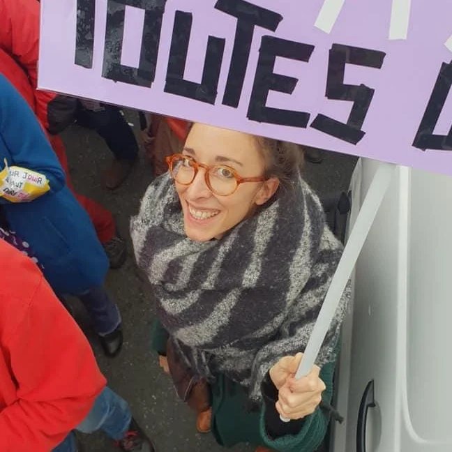 Une femme souriante portant des lunettes orange et une écharpe grise et noire, tenant une pancarte lors d'une manifestation ou d'une protestation, avec d'autres personnes en arrière-plan.