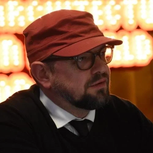 Homme avec un chapeau marron, lunettes noires, barbe et moustache, portant un chemisier blanc et une cravate noire, assis dans un lieu éclairé par un panneau lumineux rouge à l'arrière.