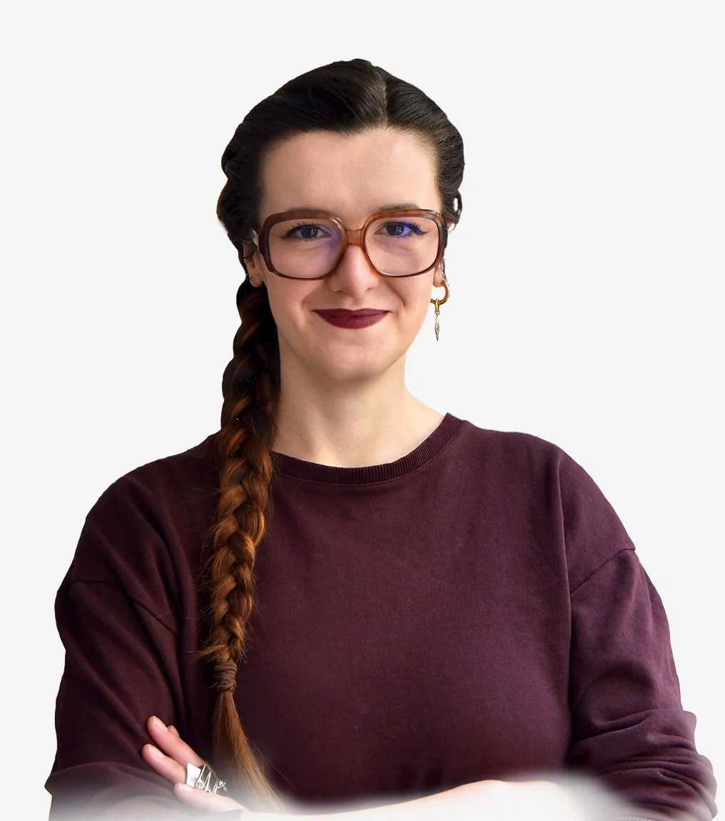 Photographie d'une femme de face souriante la bouche fermée. Elle à les cheveux bruns et très longs, jusqu'en bas du torse. Elle a les cheveux lisses et tressés sur le côté droit. Elle porte de grandes lunettes carrées et porte un pull bordeaux. 