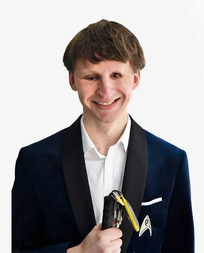 Photographie d'un homme de face, chveux châtains mi-long, tombant à haute de front. Il se tient face à la caméra avec sa canne jaune à la main droite. Il souri et est habillé d'une chemise blanche et d'une veste de costume noire.