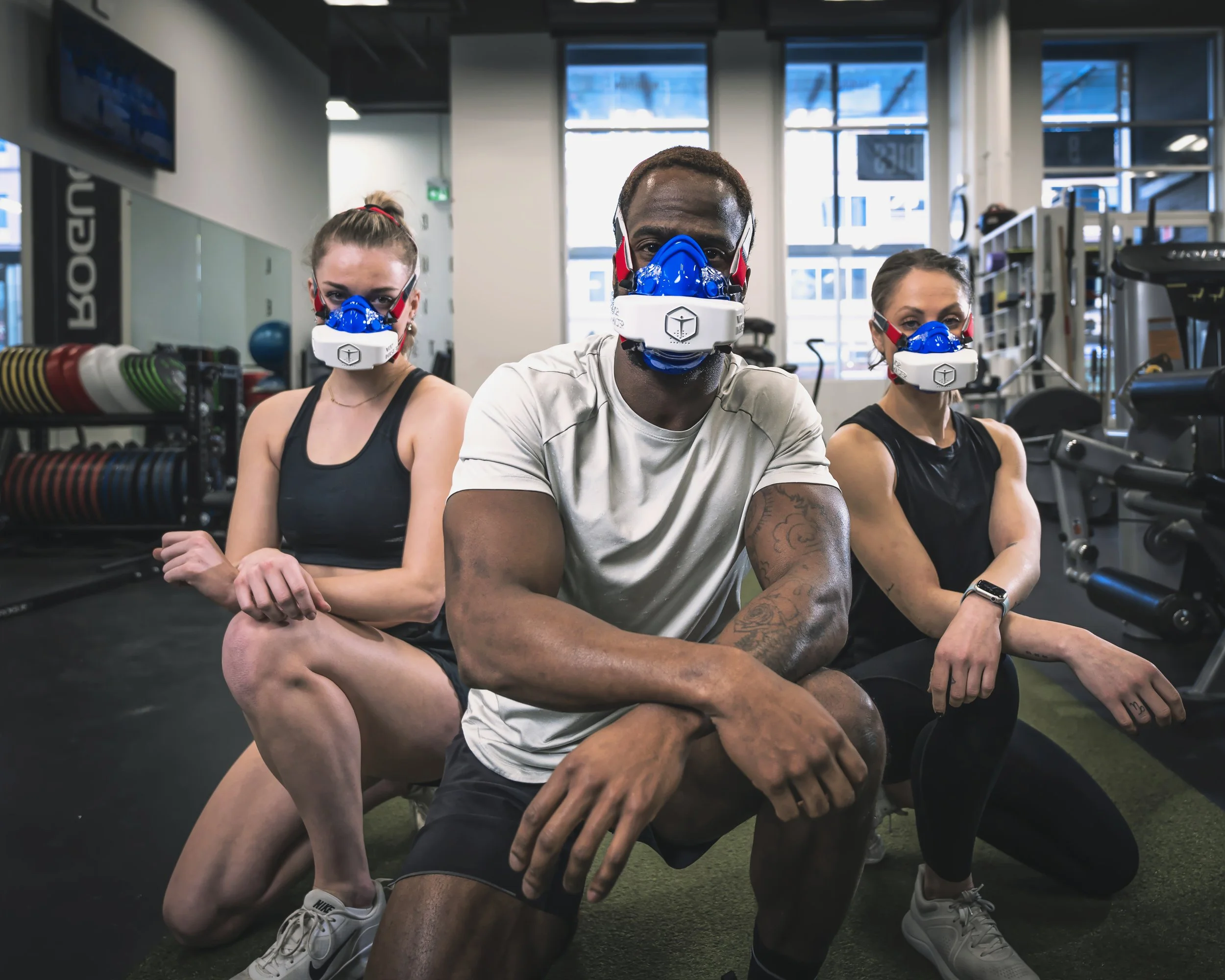 Three people in a gym wearing protective masks with blue filters, sitting on the floor. The gym has workout equipment and weight racks in the background.