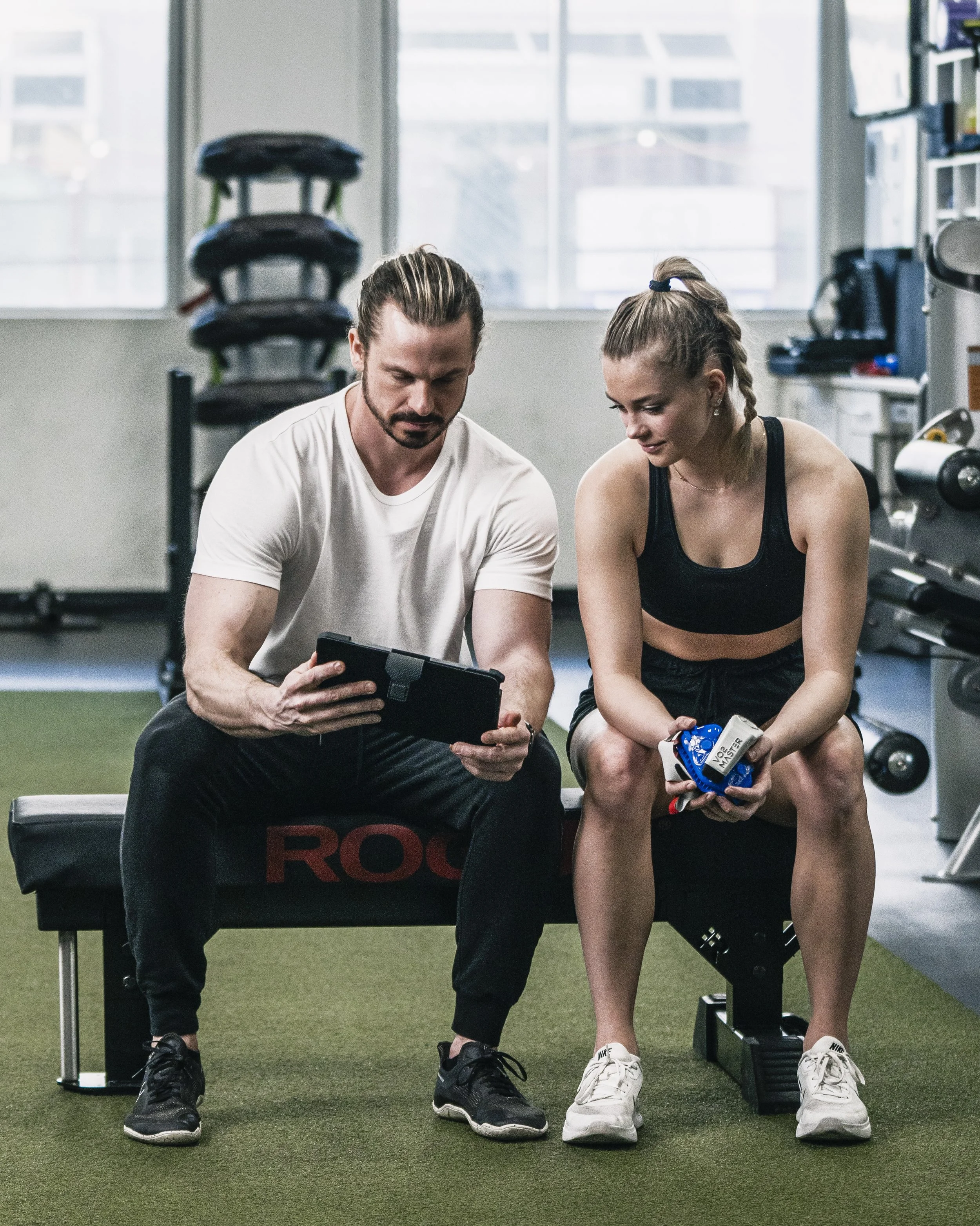 A man and a woman sitting on a bench in a gym, looking at a tablet and a fitness tracker respectively.
