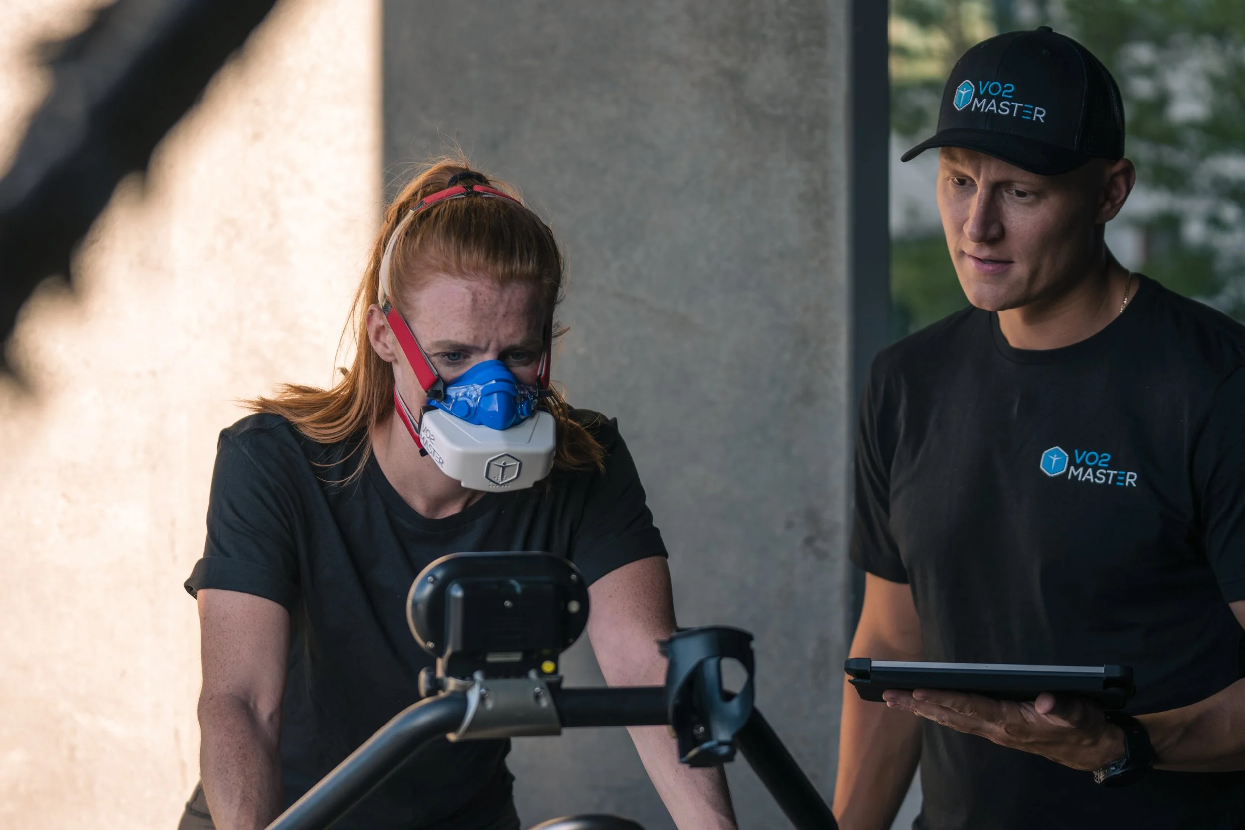 A woman wearing a breathing mask on an exercise bike, with a trainer standing beside her holding a tablet, in a gym or fitness studio.