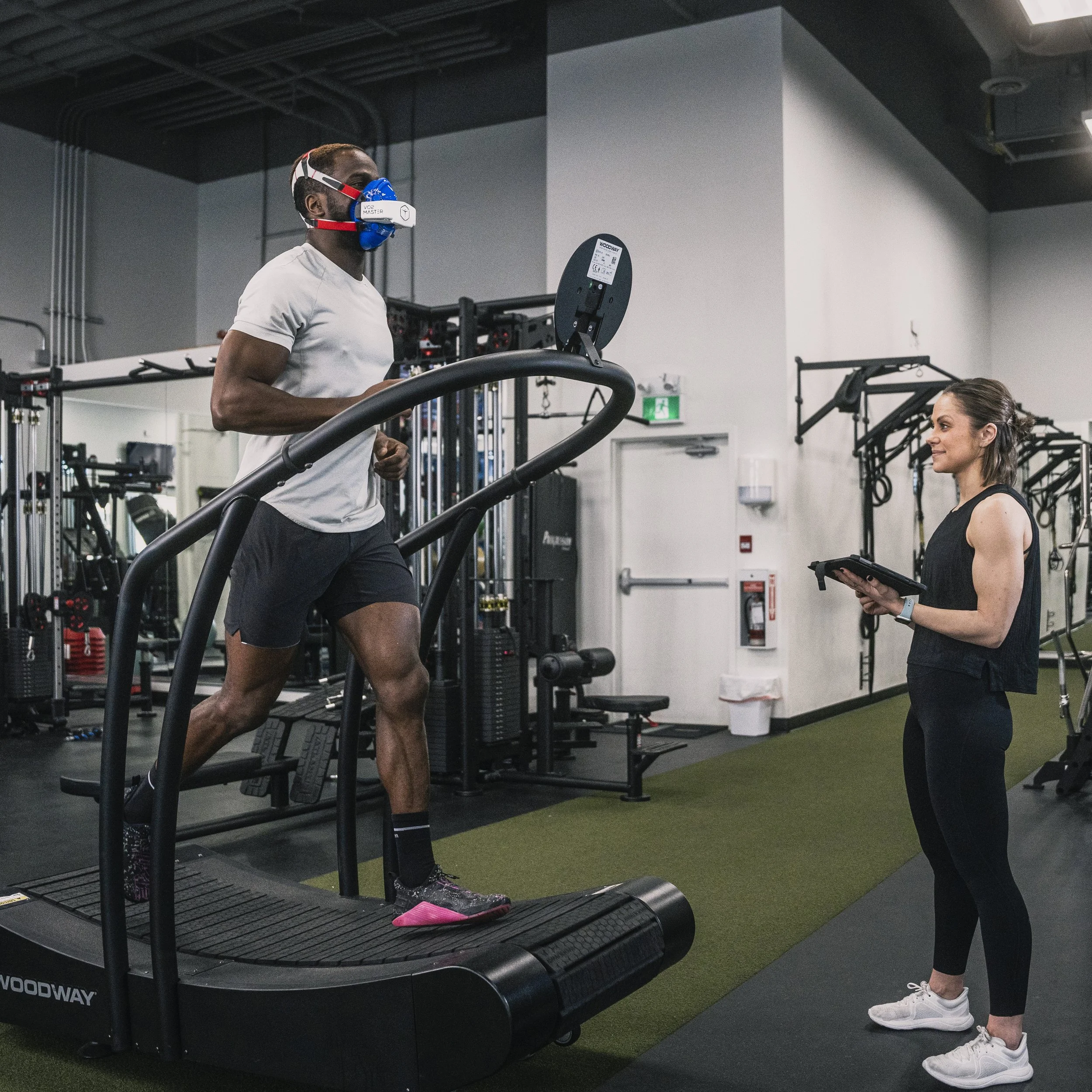 A man running on a treadmill with a face mask for training, while a woman holding a clipboard supervises in a gym.