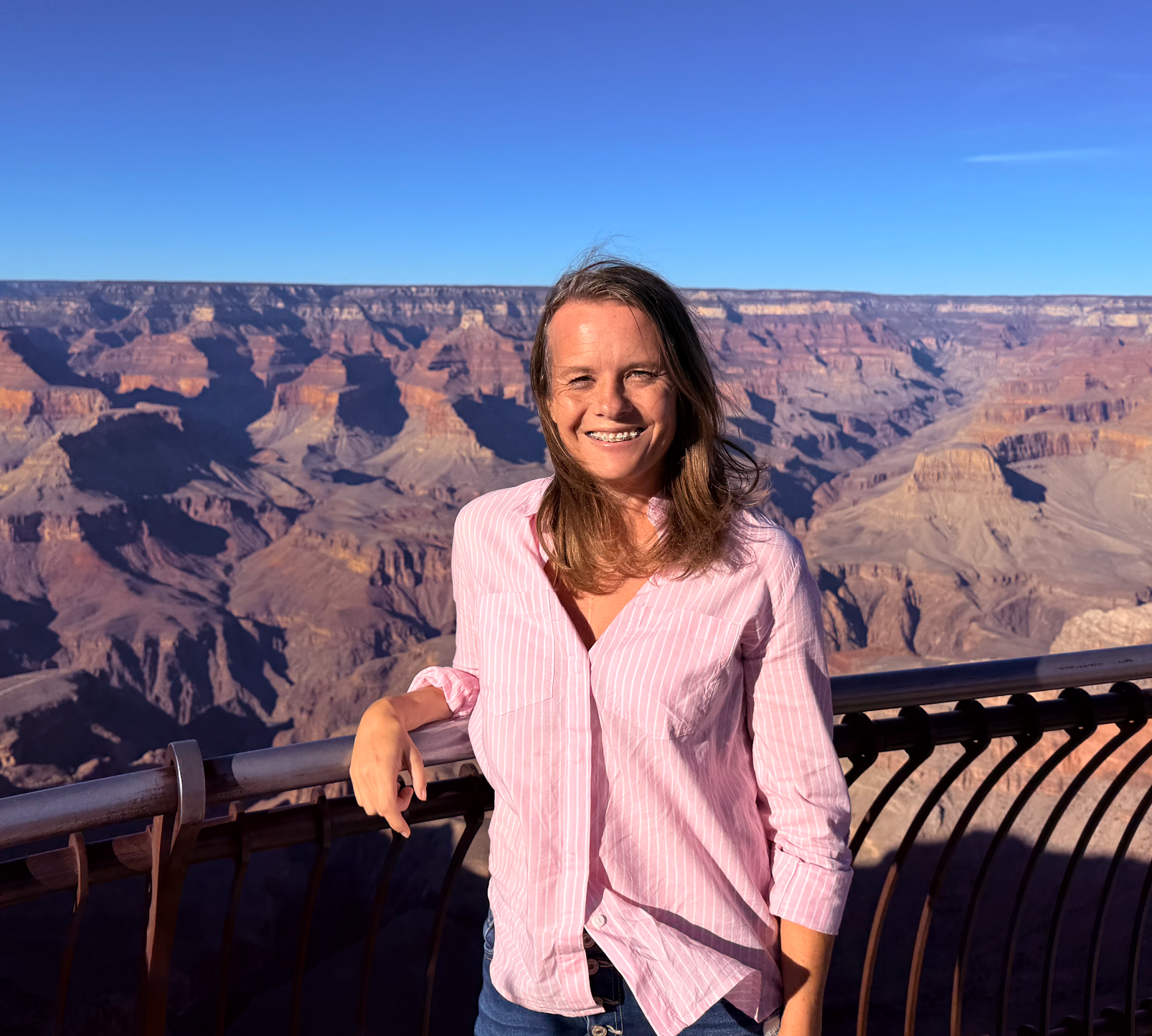 A woman smiling at the camera while standing at the Grand Canyon viewing platform with the canyon and blue sky in the background.