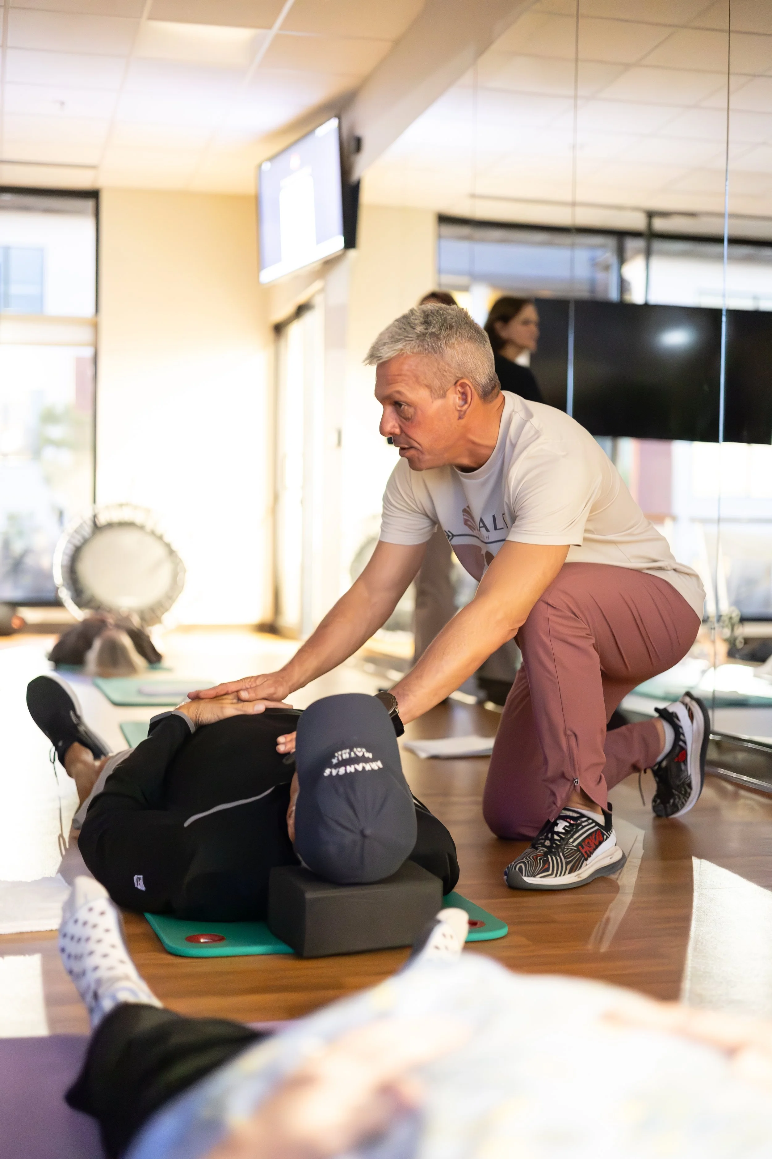 A man providing Breathwork training to a woman lying on a mat, with other participants practicing in the background, inside a well-lit room with fitness equipment.