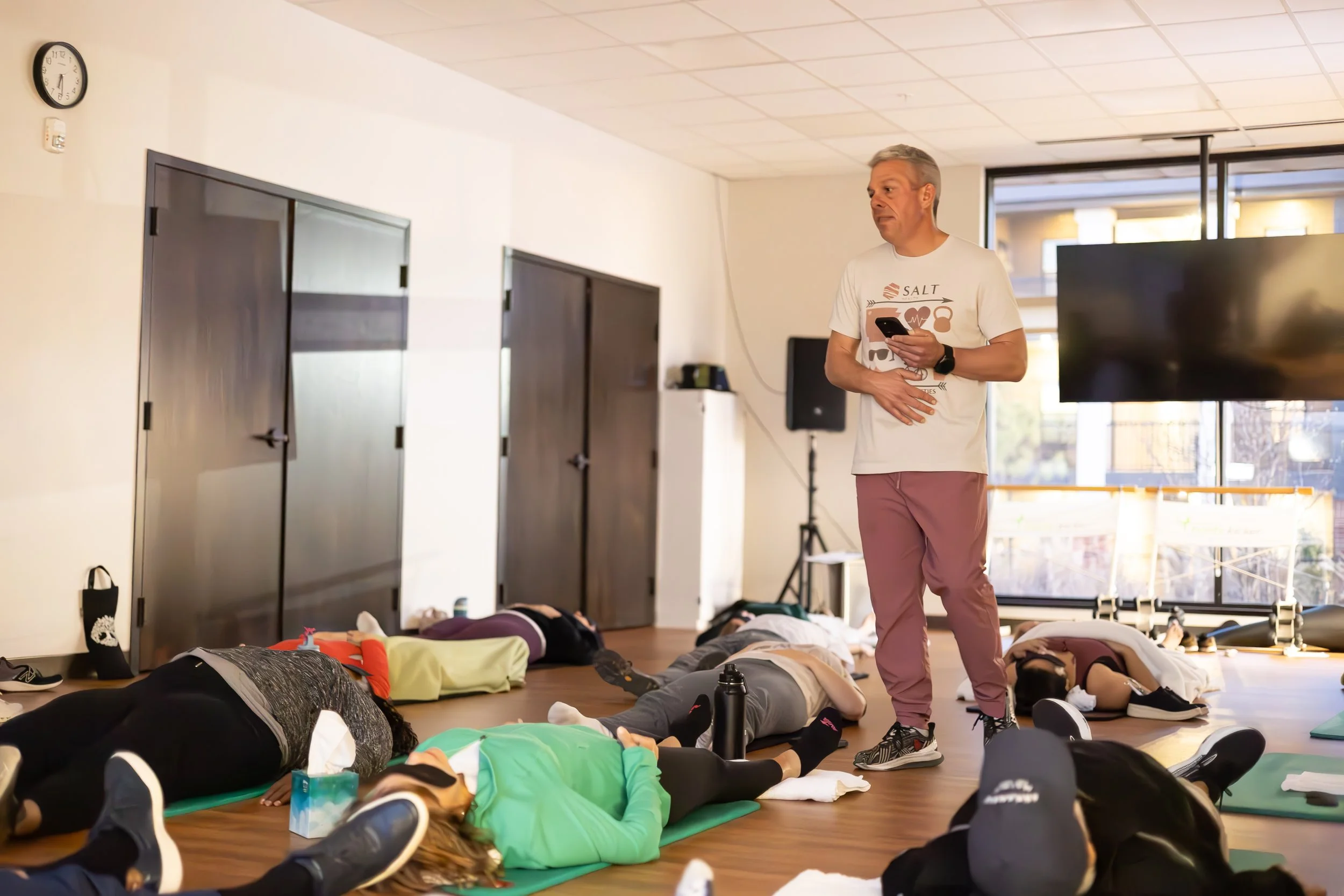 Instructor standing in front of a breath work class, demonstrating safety techniques, with students lying on mats practicing in a bright studio.