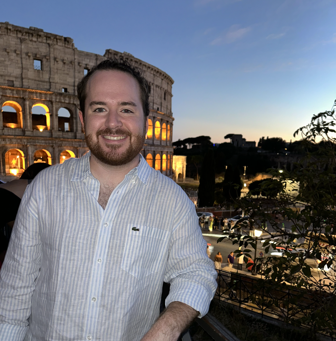 A man smiling in front of the Colosseum at dusk in Rome, Italy.