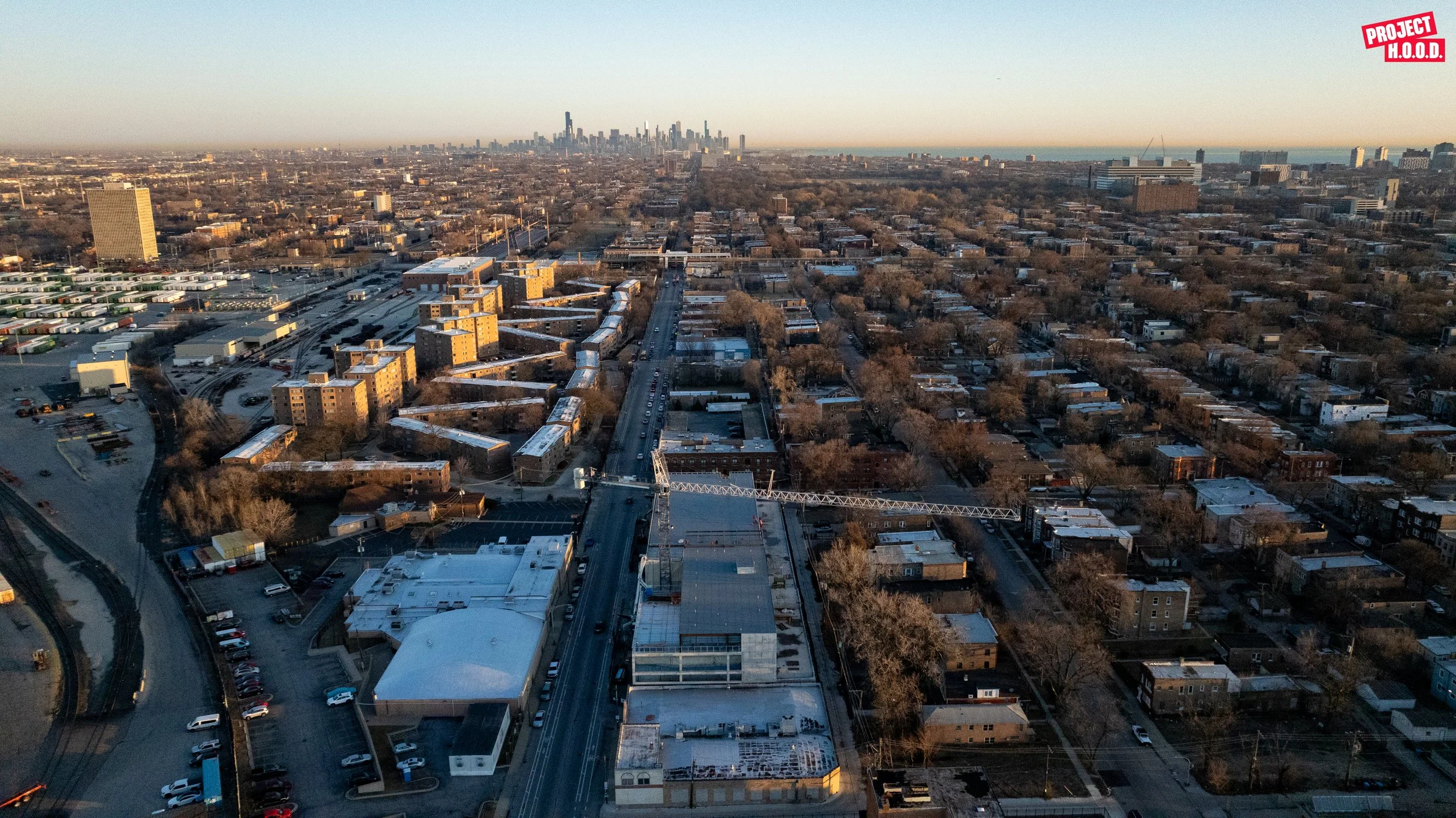 Aerial view of a cityscape showing streets, buildings, and residential neighborhoods with a downtown skyline in the distance, taken during sunset.